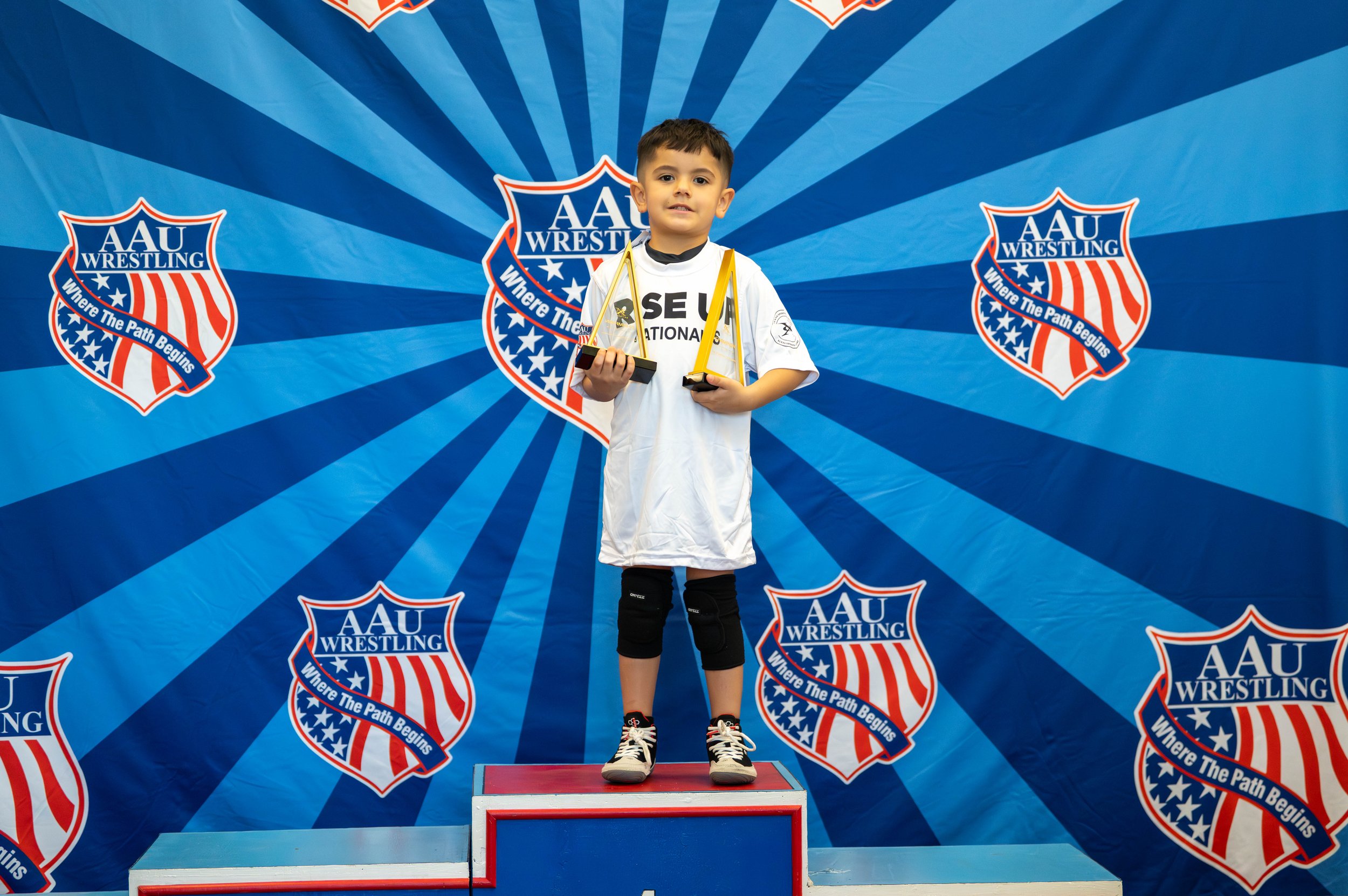 Young boy standing on a winner's podium holding two trophies, with a backdrop featuring the AAU Wrestling logo and the slogan "Where the Path Begins."