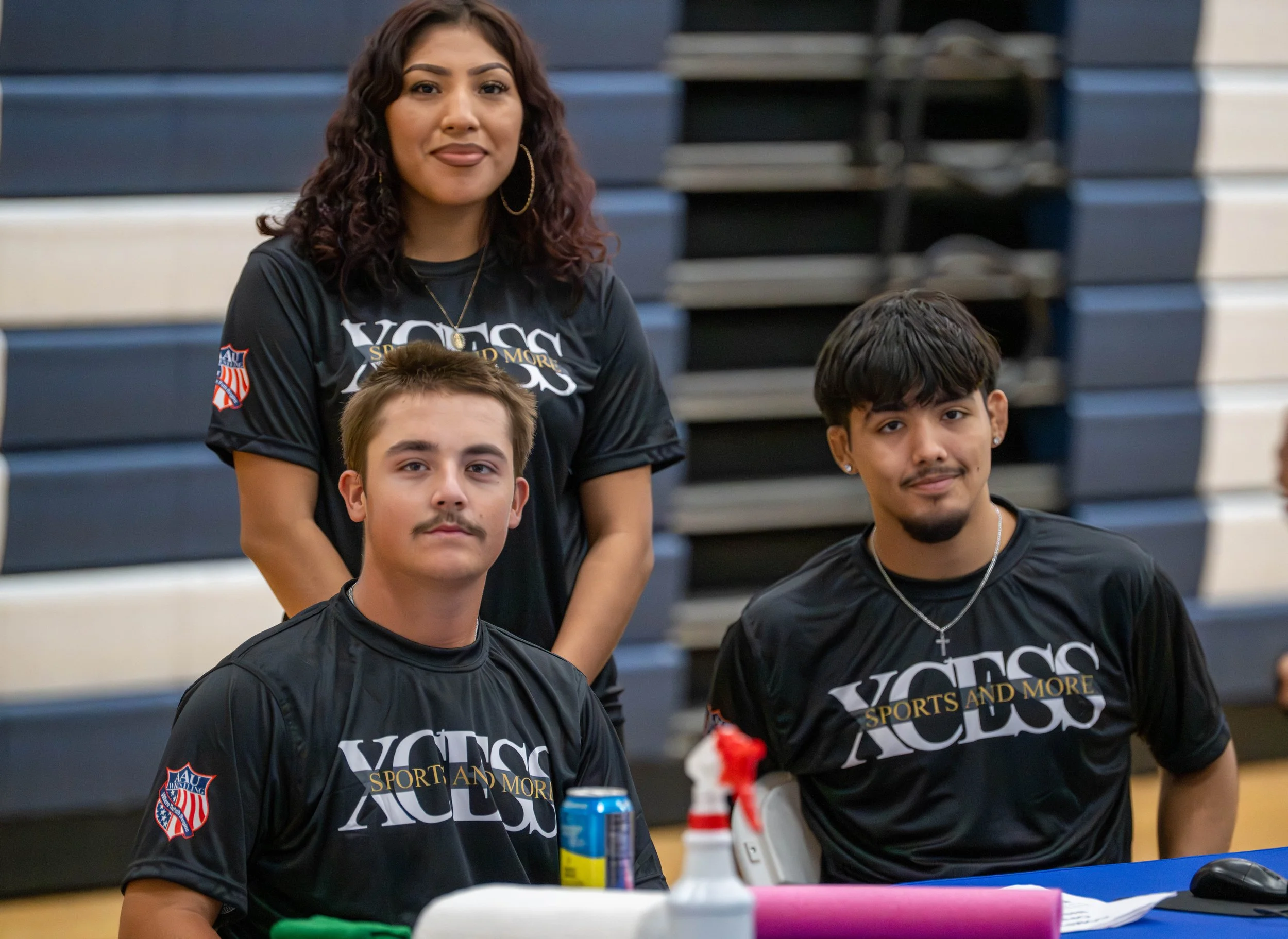 A woman and two young men sitting and standing at a table in a gymnasium. They are all wearing black sports T-shirts with 'VOSS SPORTS AND MORE' written on them. The woman has curly dark hair, and the young men have short hair, one with a mustache an