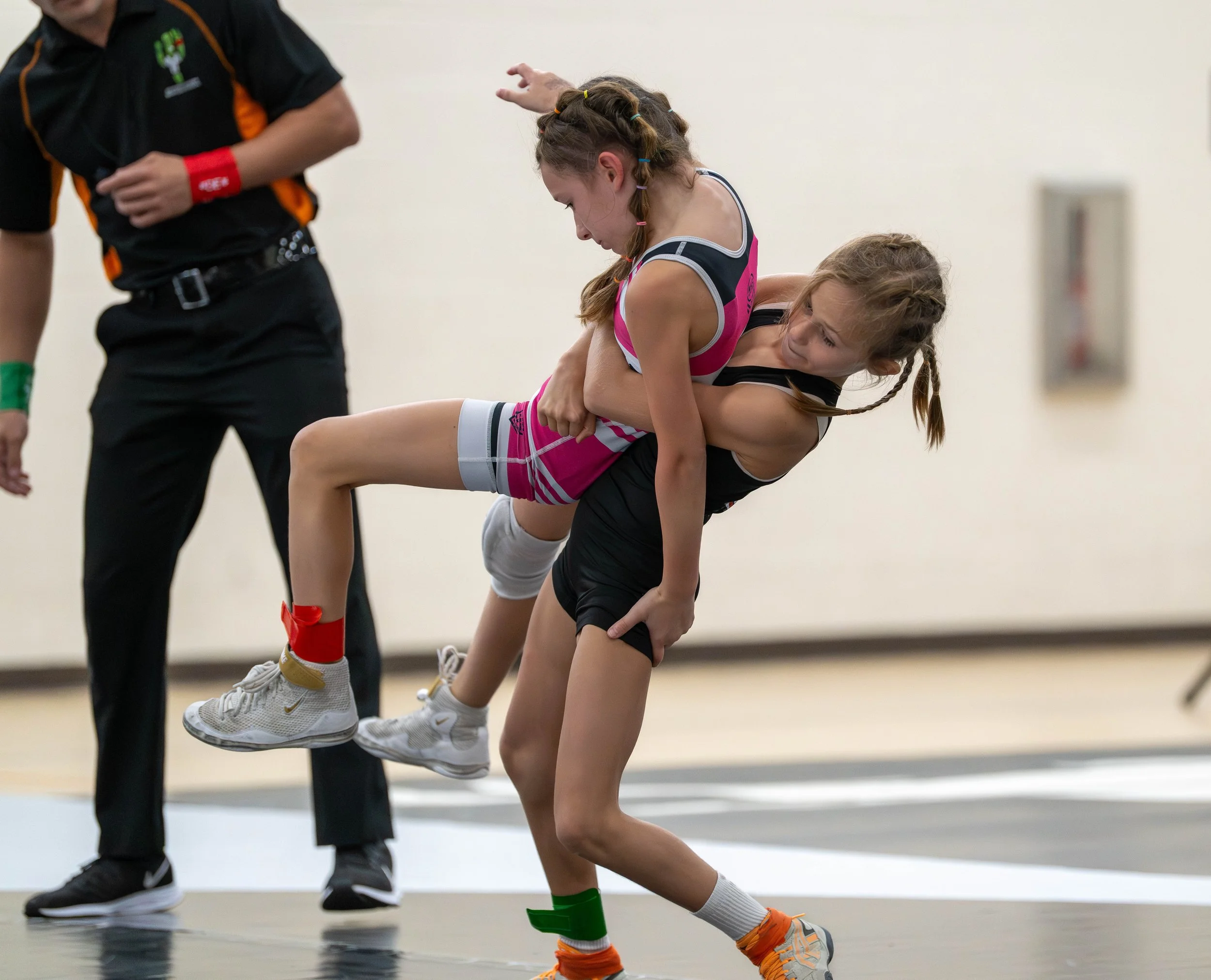 Two young girls wrestling on a mat, with one girl in black and the other in pink and gray, and an official standing nearby.