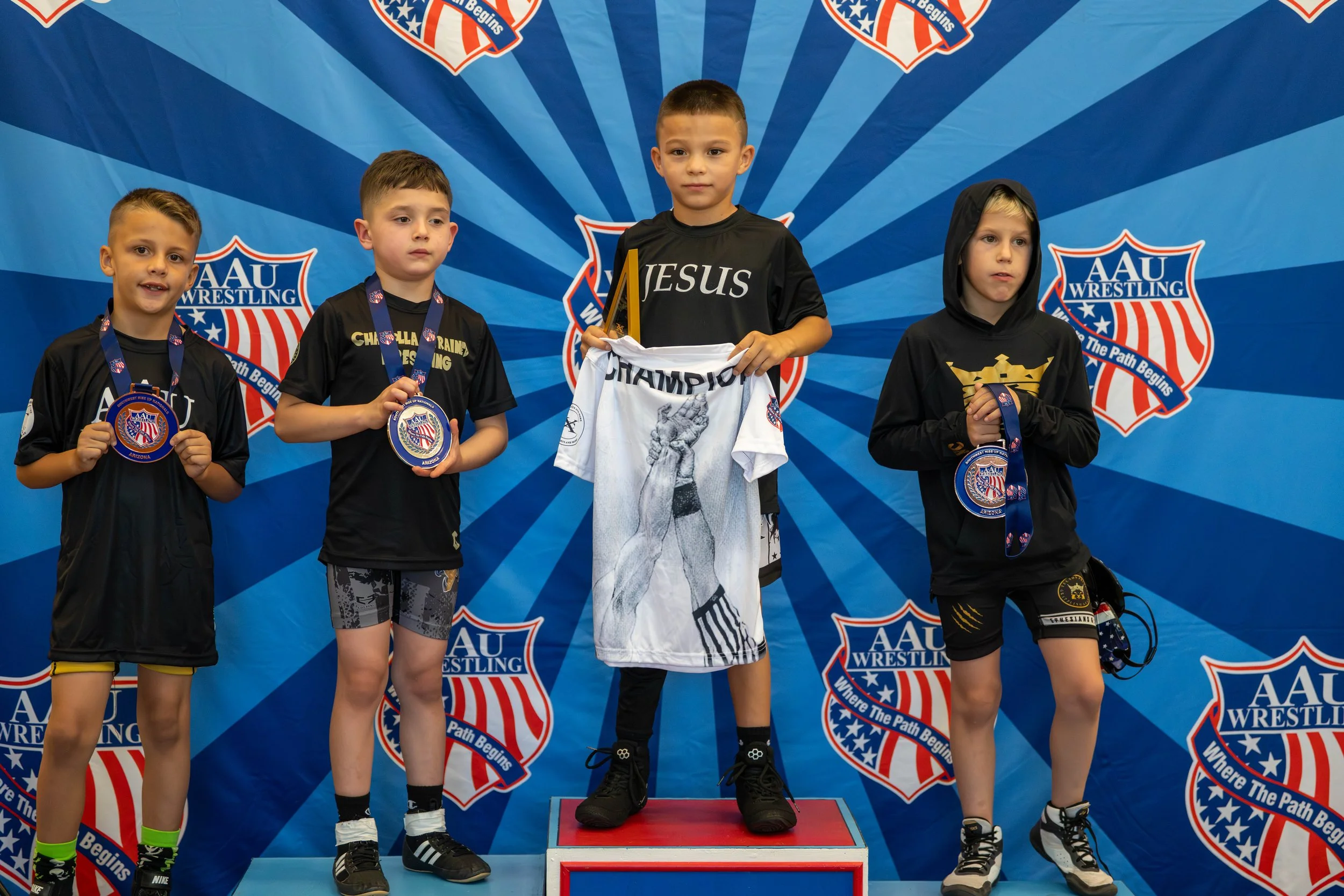 Young boys standing on a podium holding medals and a shirt with the words 'JESUS CHAMPION' at an AAU wrestling event.