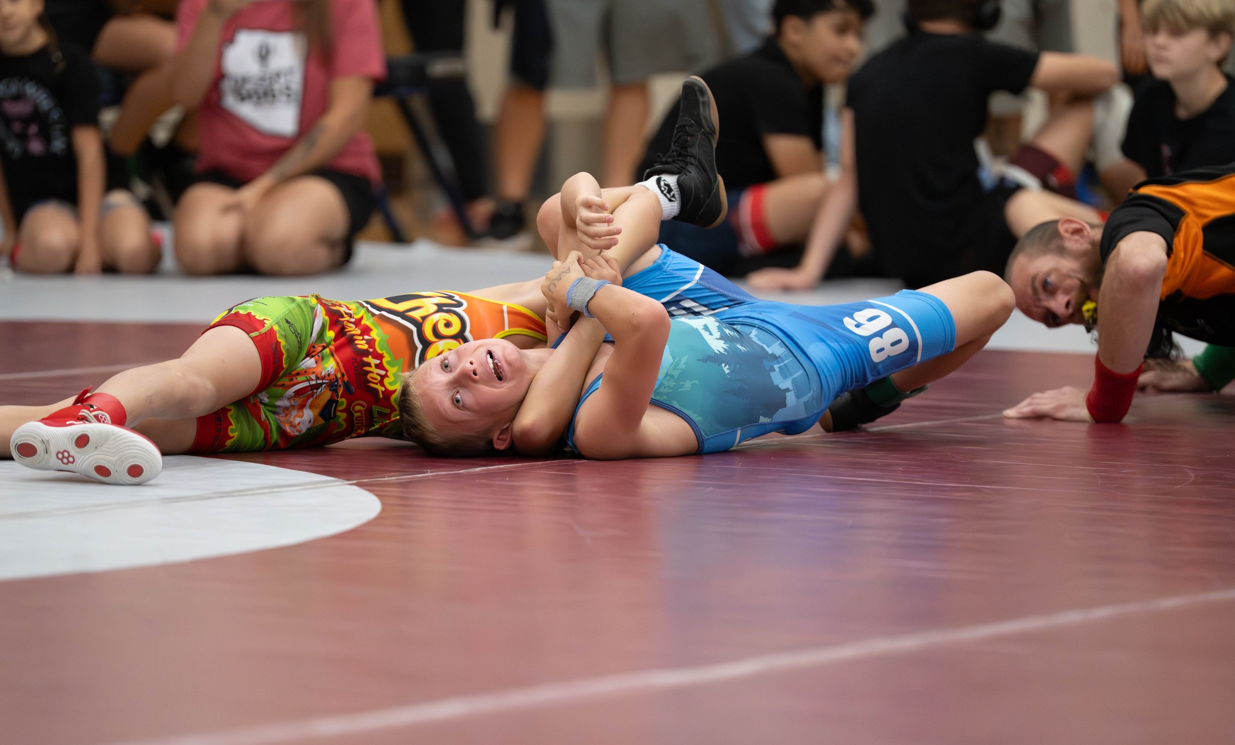 Two young wrestlers on a mat, one in a colorful shirt and the other in a blue singlet, grapple during a match, with at least one referee and several children sitting in the background.