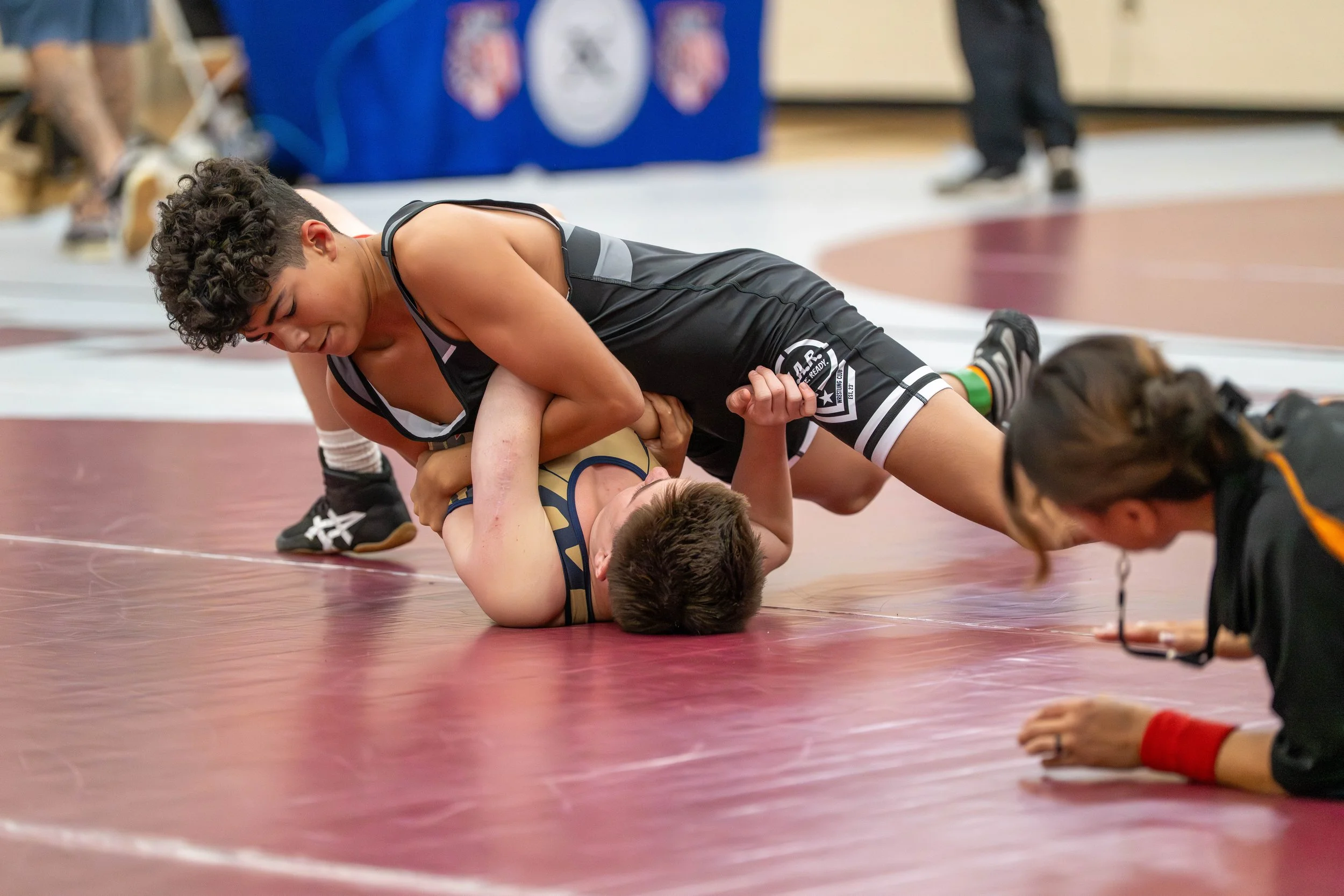 Two male wrestlers are engaged in a wrestling match on a maroon mat, with one wrestler on top and the other on the ground, while a referee looks on. The scene appears to be in a gymnasium or sports arena.