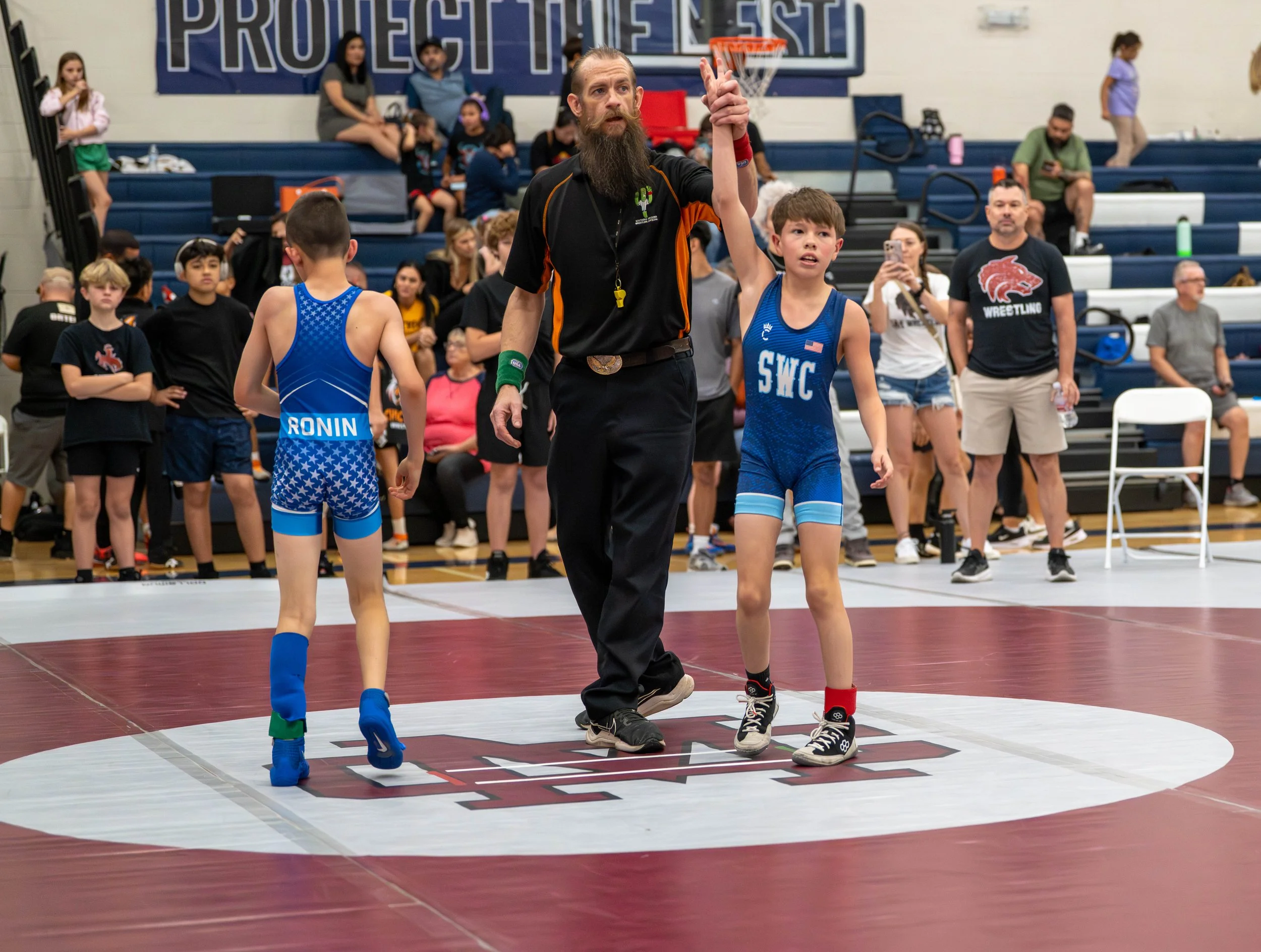 A youth wrestling match is concluded with a referee raising the winner's hand. The winner and the other competitor are young boys wearing wrestling singlets, with the victor's singlet marked 'SWC'. The match takes place in a gymnasium filled with spe