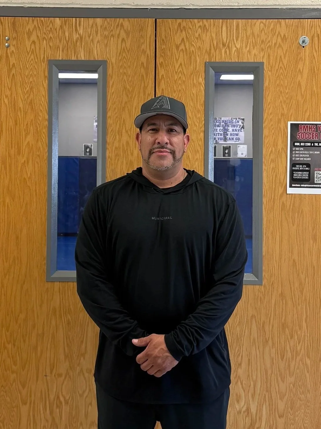 A man with a goatee and mustache stands in front of wooden double doors with narrow vertical windows, wearing a black hoodie and a gray baseball cap.