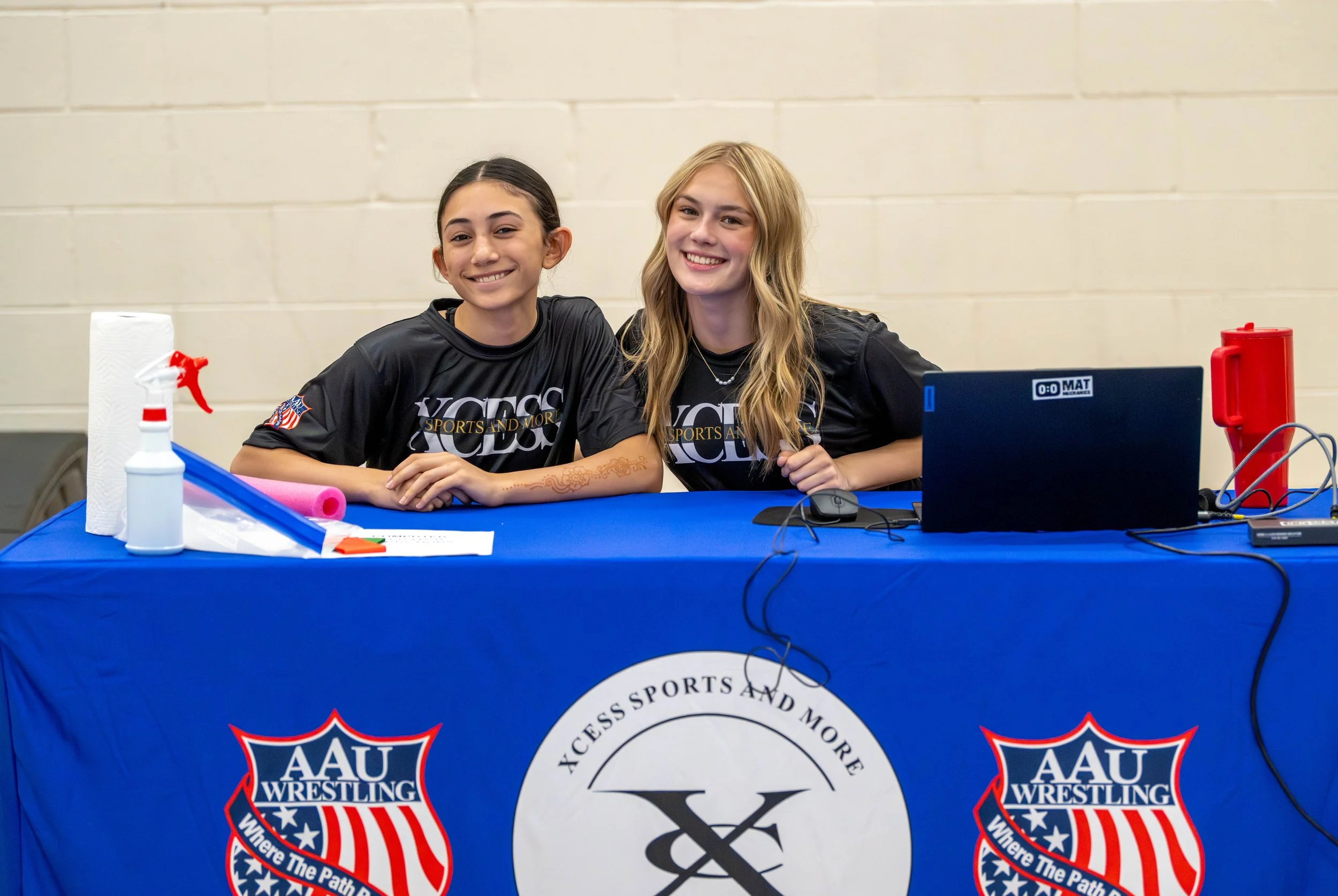 Two young women sitting at a table with AAU Wrestling banner, smiling at the camera, in a gymnasium.