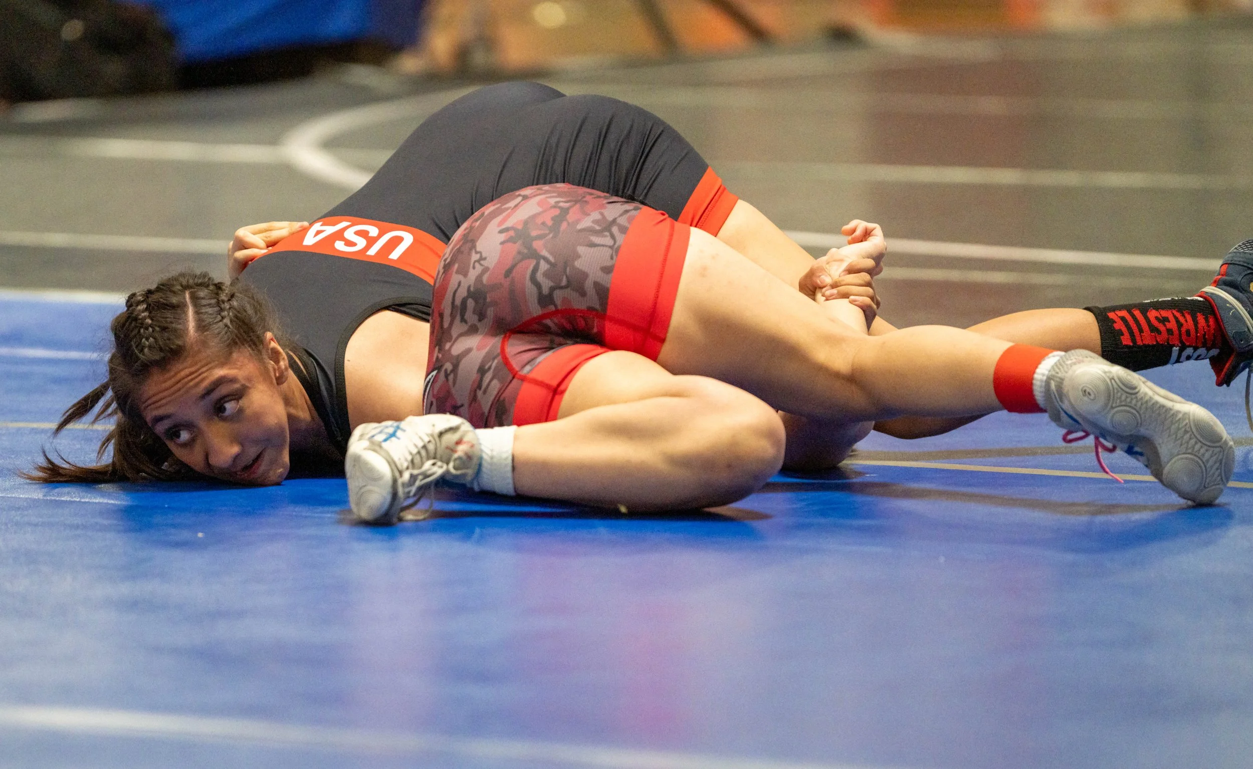 Two female wrestlers on a blue wrestling mat during a match, with one in a black and red USA uniform and the other in camouflage red and black shorts.