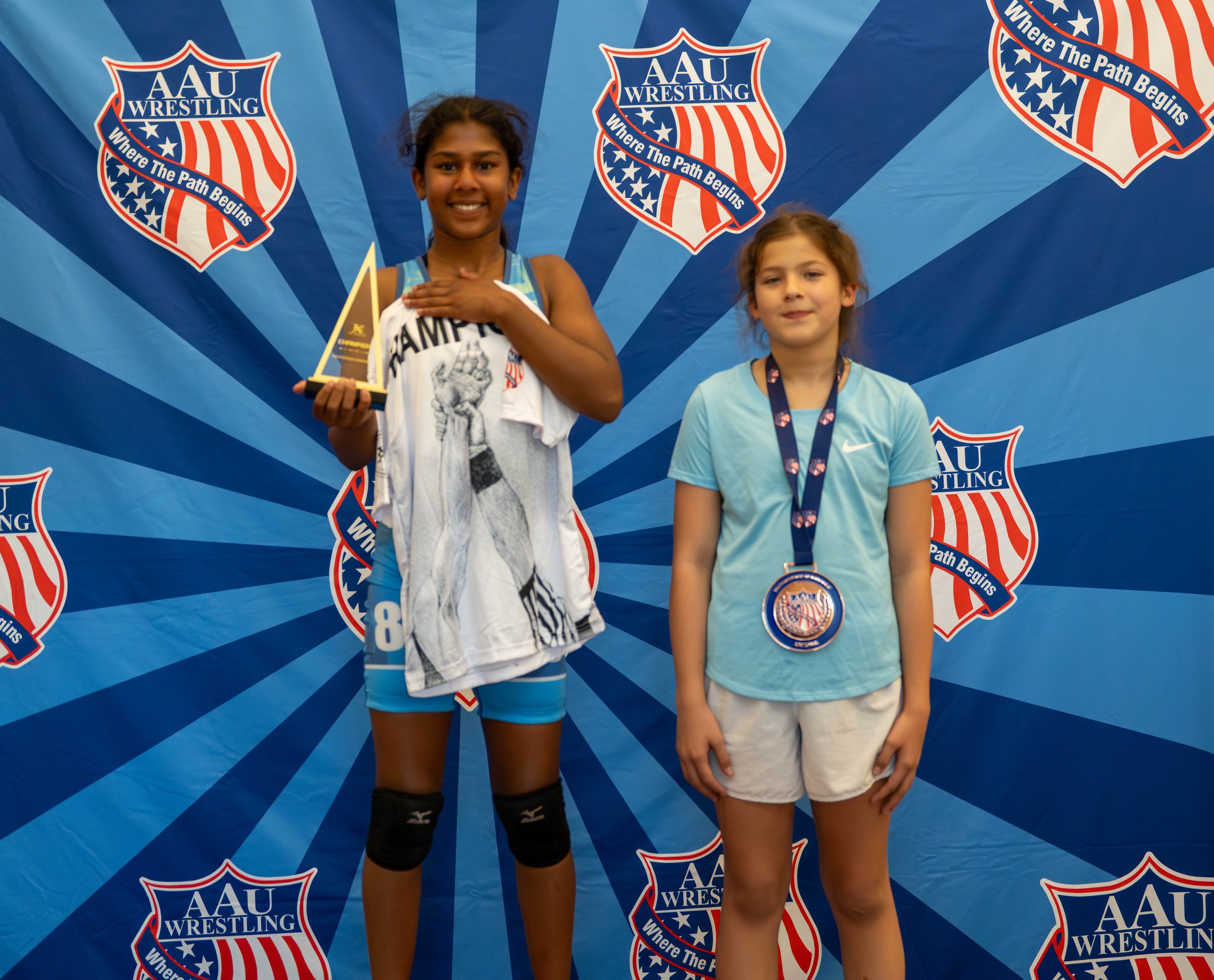 Two young girls standing on a winners' podium at an AAU Wrestling event, with a backdrop displaying the AAU Wrestling logo and the slogan 'Where The Path Begins'. The girl on the left is holding a trophy and wearing a medal, while the girl on the rig