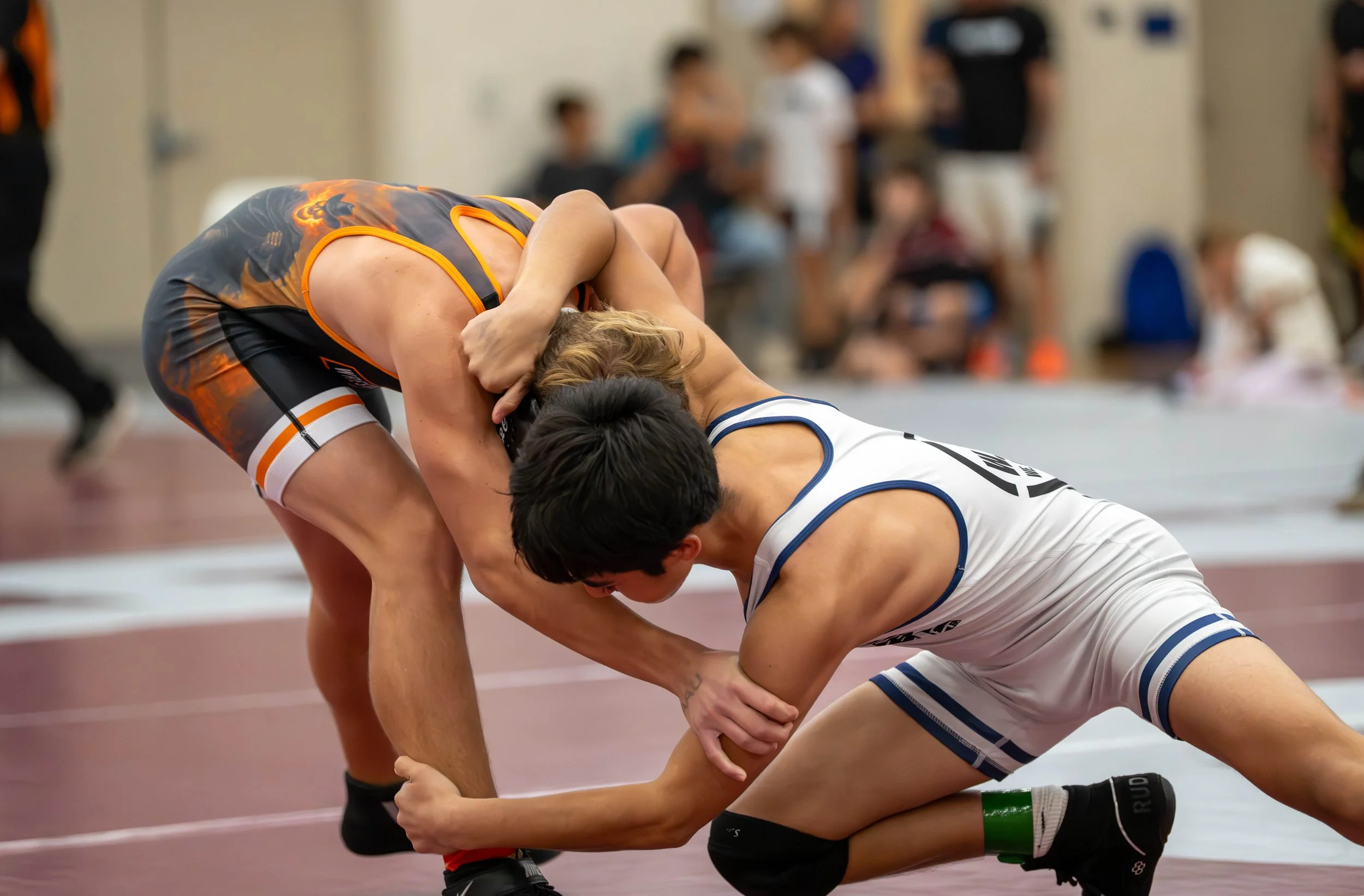 Two wrestlers, one in a black and orange singlet and the other in a white singlet with blue trim, are engaged in a match on a wrestling mat indoors. The wrestler in white is on one knee, gripping the legs of the wrestler in black and orange, who is b