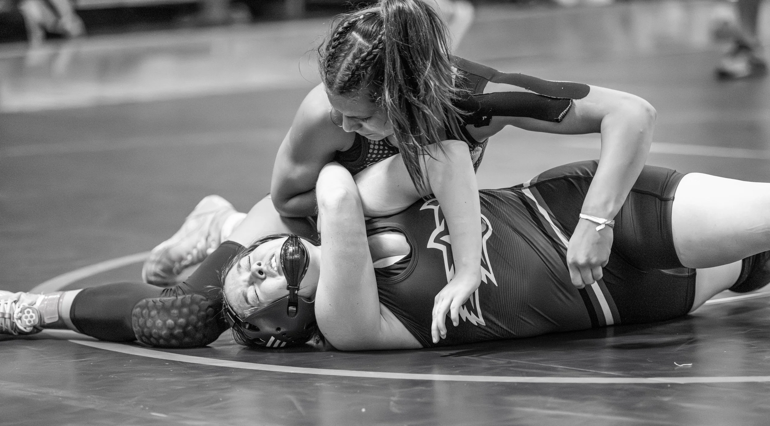 Two female wrestlers engaged in a match on a wrestling mat, one on top of the other, with one wearing a protective headgear and athletic gear.