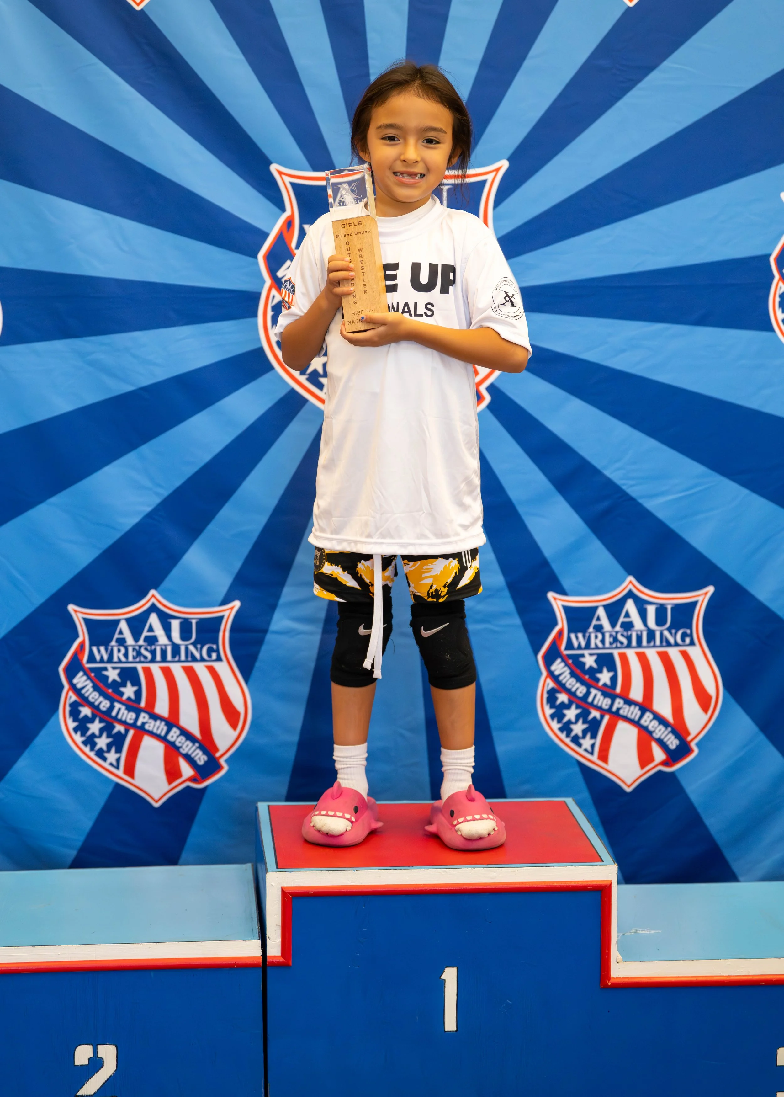 Young girl standing on a winners' podium holding a trophy, wearing a wrestling jersey, shorts, knee pads, and pink slippers, with a blue backdrop featuring AAU Wrestling logos.