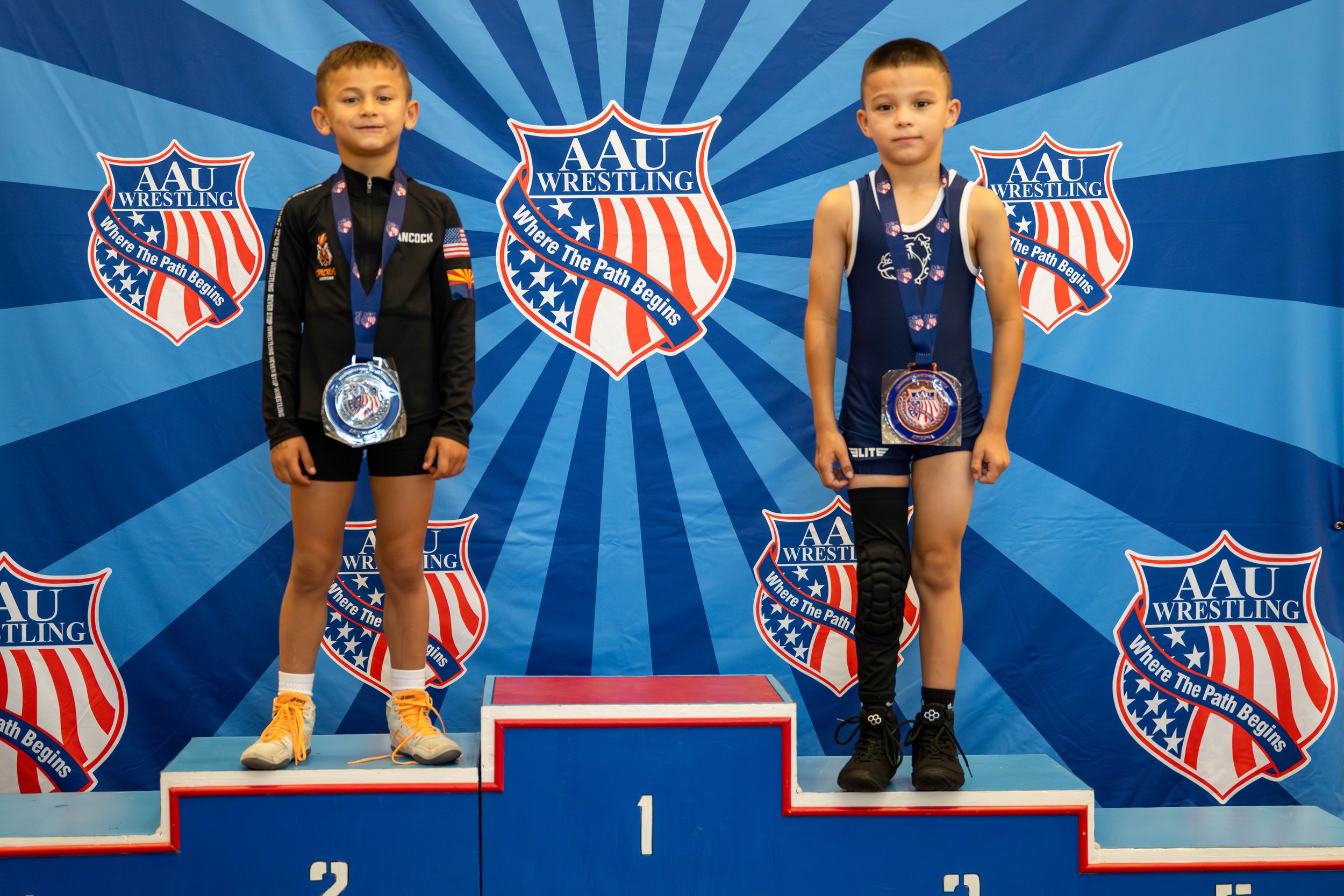 Two young boys in wrestling uniforms standing on a winners' podium with medals, with a backdrop featuring the AAU Wrestling logo and the slogan 'Where The Path Begins'.