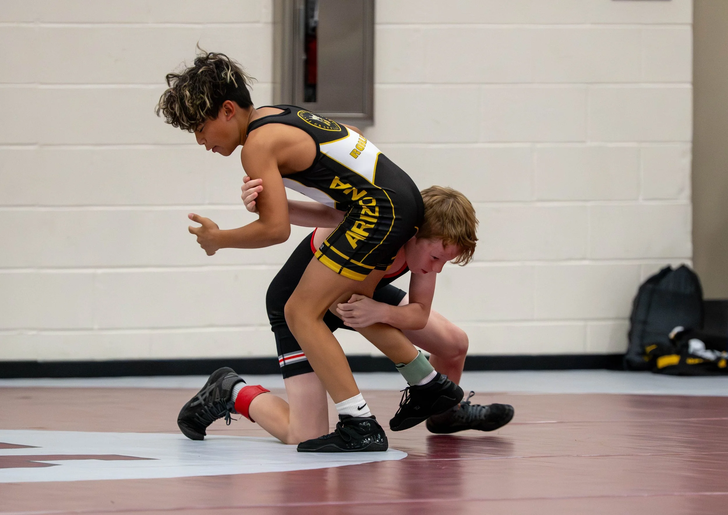 Two young wrestlers in black and yellow singlets are grappling on a wrestling mat in a gym. One wrestler is in a crouch holding the other wrestler in a double leg takedown position.