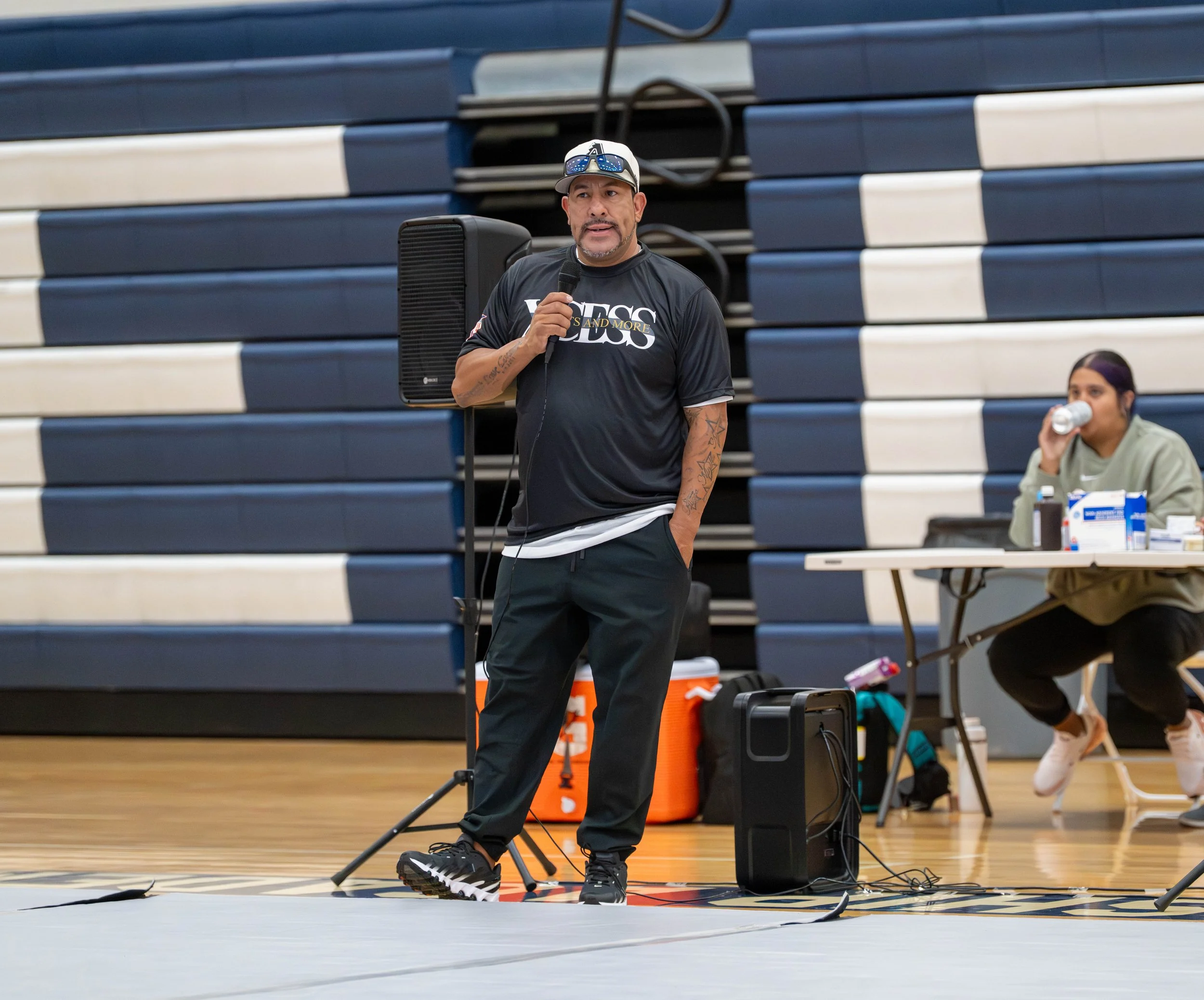 A man standing in a gymnasium, speaking into a microphone, with a woman sitting at a table drinking water in the background.