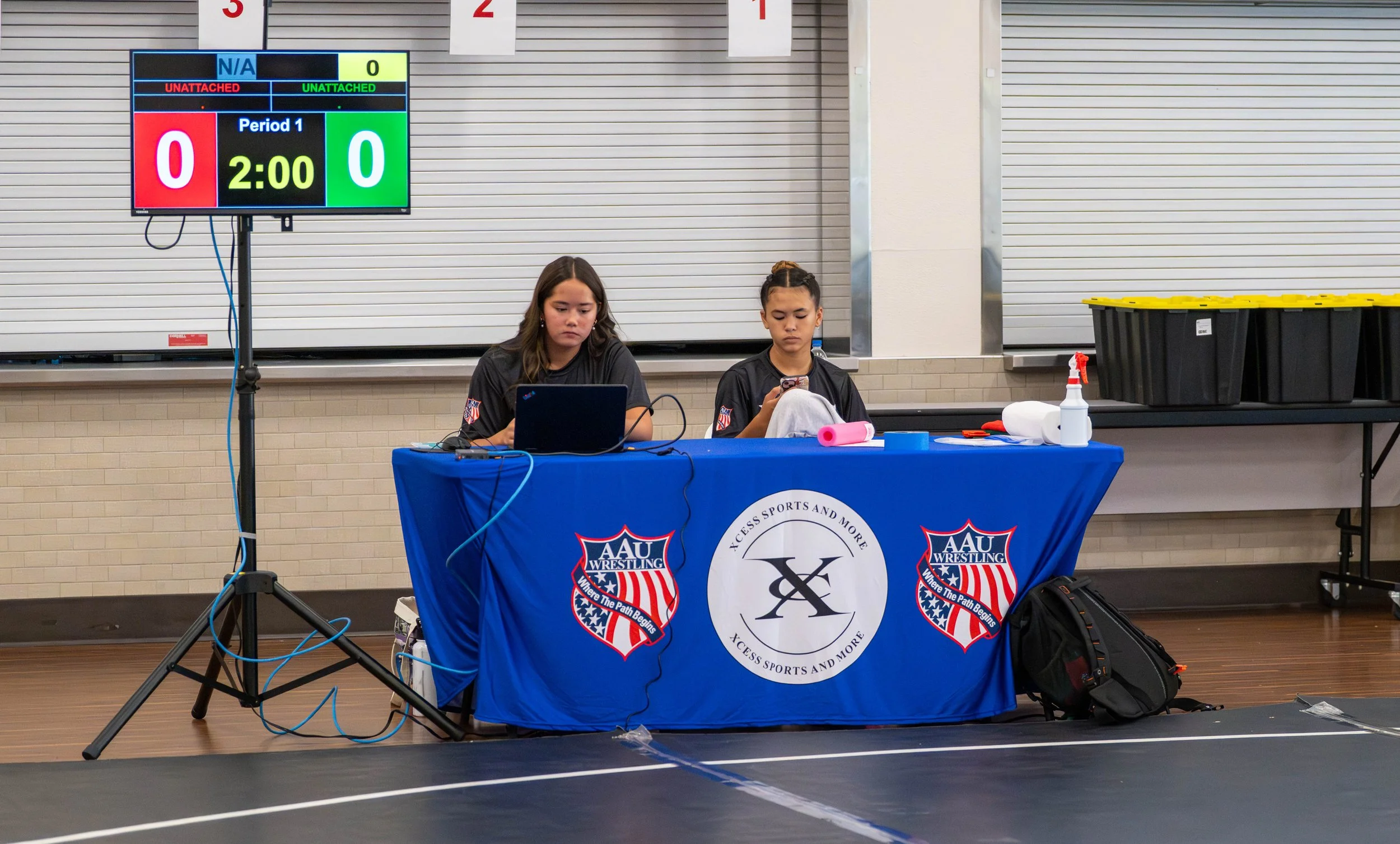Two young female wrestlers sit at a table with laptops in a gymnasium during a wrestling event. The table is draped with a blue cloth featuring the AAU Wrestling logo and a circular logo in the center. Behind them, a scoreboard displays a tied score 