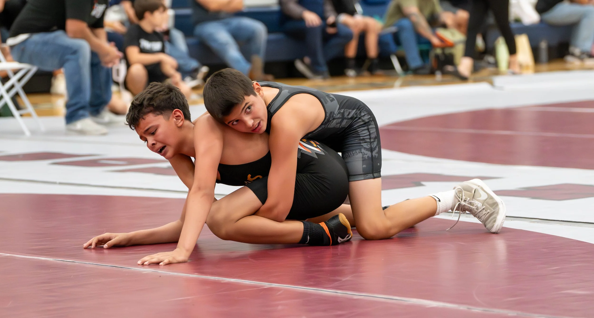 Two young boys wrestling on a red mat during a match, with spectators seated in the background.