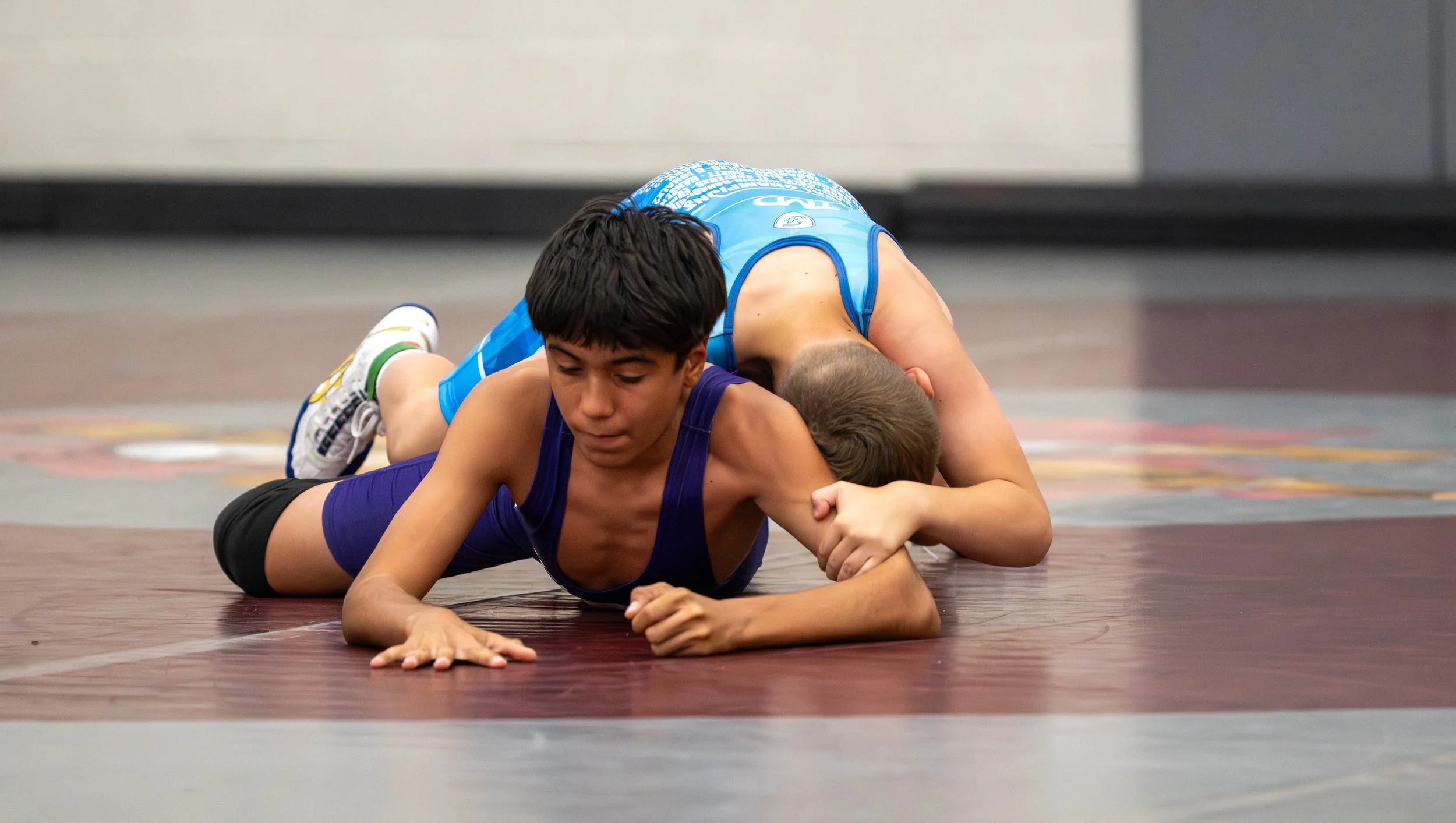 Two young boys wrestling on a mat during a match, with one in a blue singlet and the other in a purple singlet, both focused on the sport.