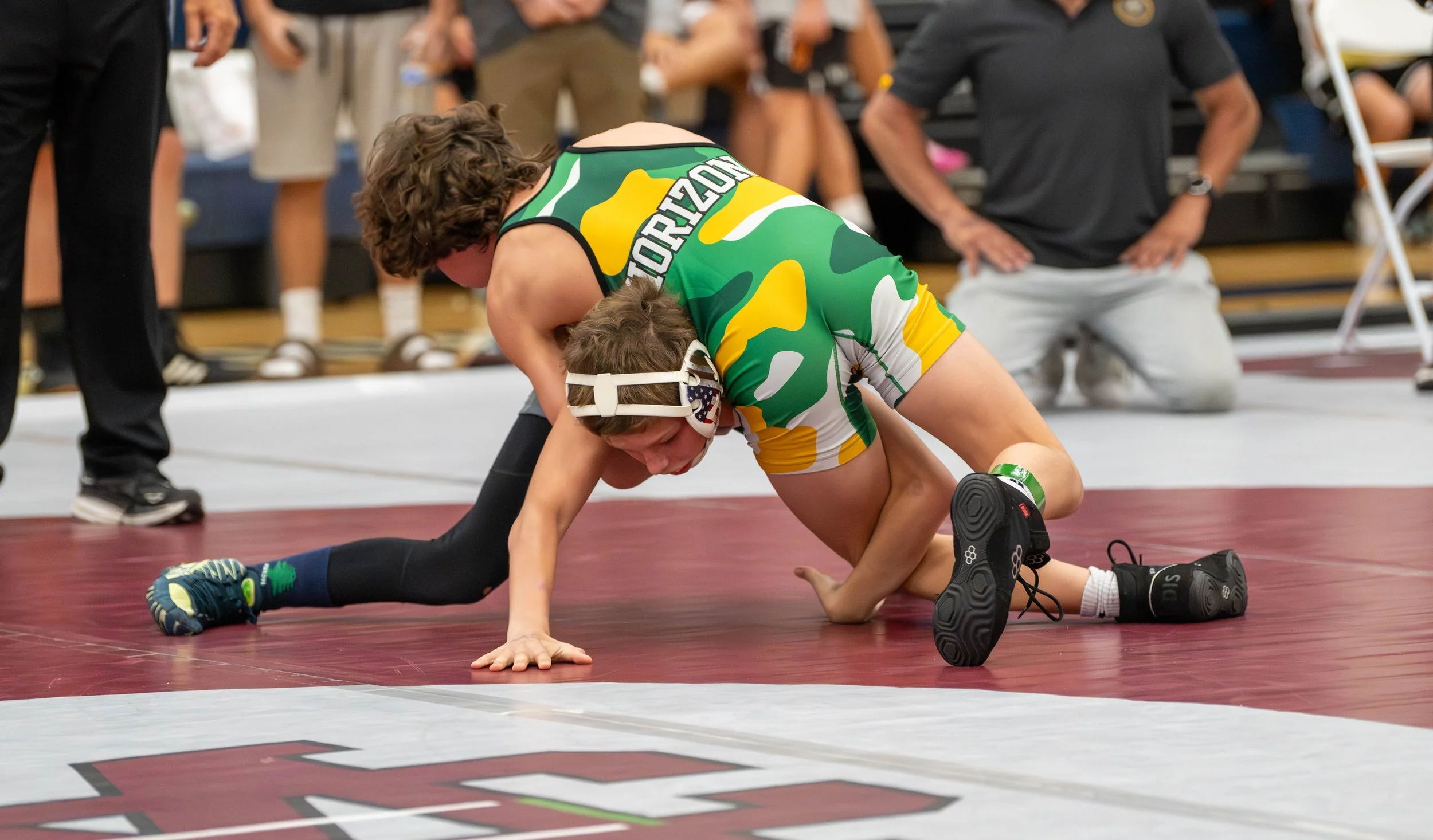 Two young wrestlers compete on the mat during a wrestling match, with one wearing a camouflage singlet and the other wearing black and green gear, while an official and spectators observe in the background.