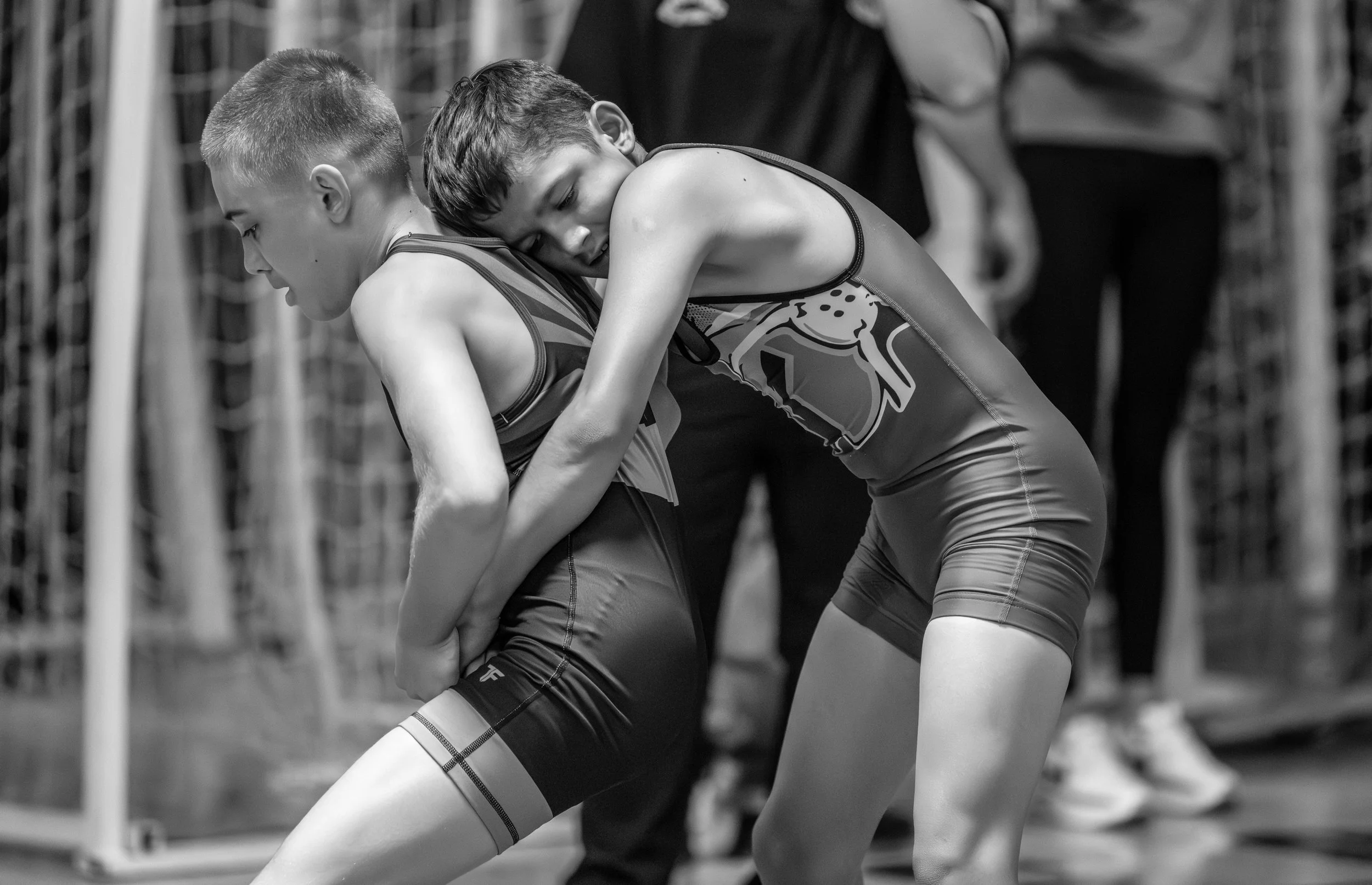 Two young wrestlers grapple during a match, one in a sleeveless sports uniform leaning forward, the other in a form-fitting singlet leaning on her opponent with a focused expression, black and white photo.