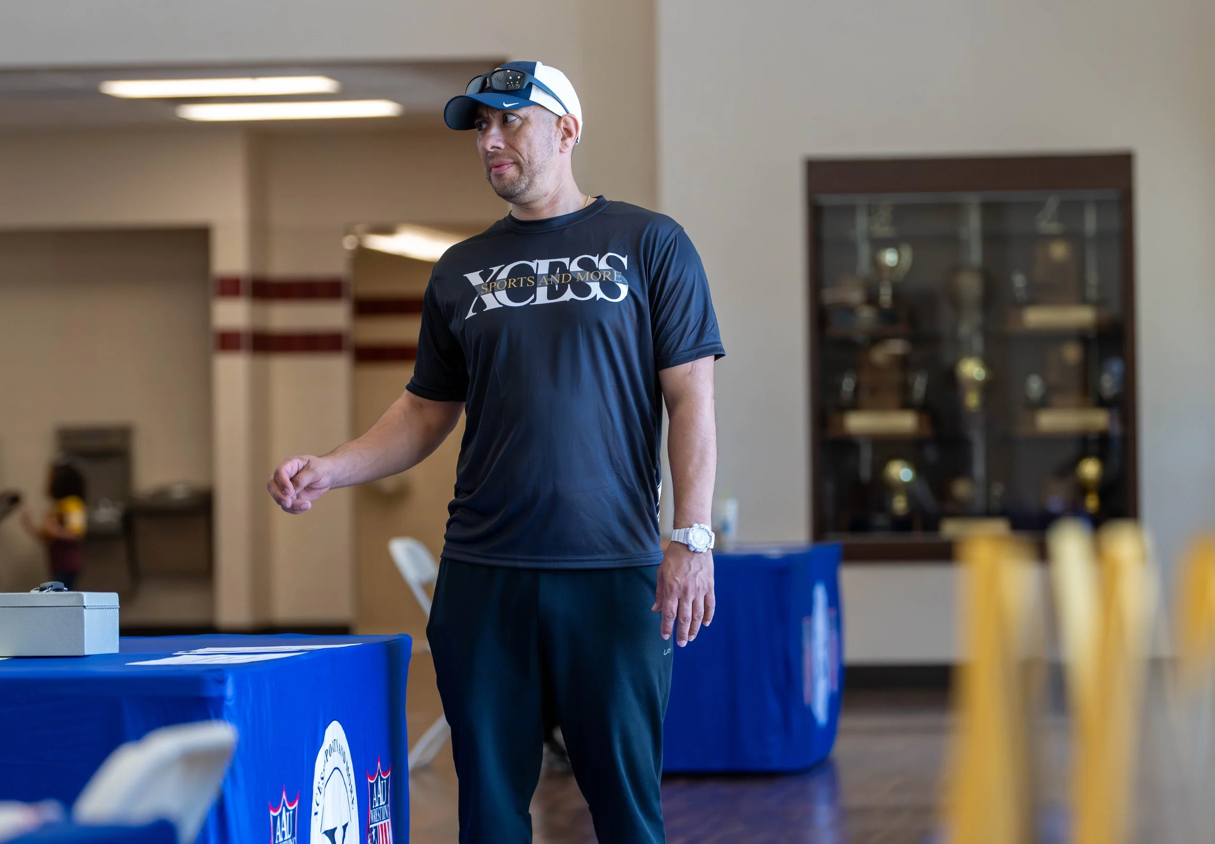 A man wearing a black t-shirt with the words 'VOLS SPORTS AND MORE' printed on it, a blue cap, and sunglasses on his head, standing indoors near a table covered with a blue tablecloth. The background includes a display cabinet with trophies and some 