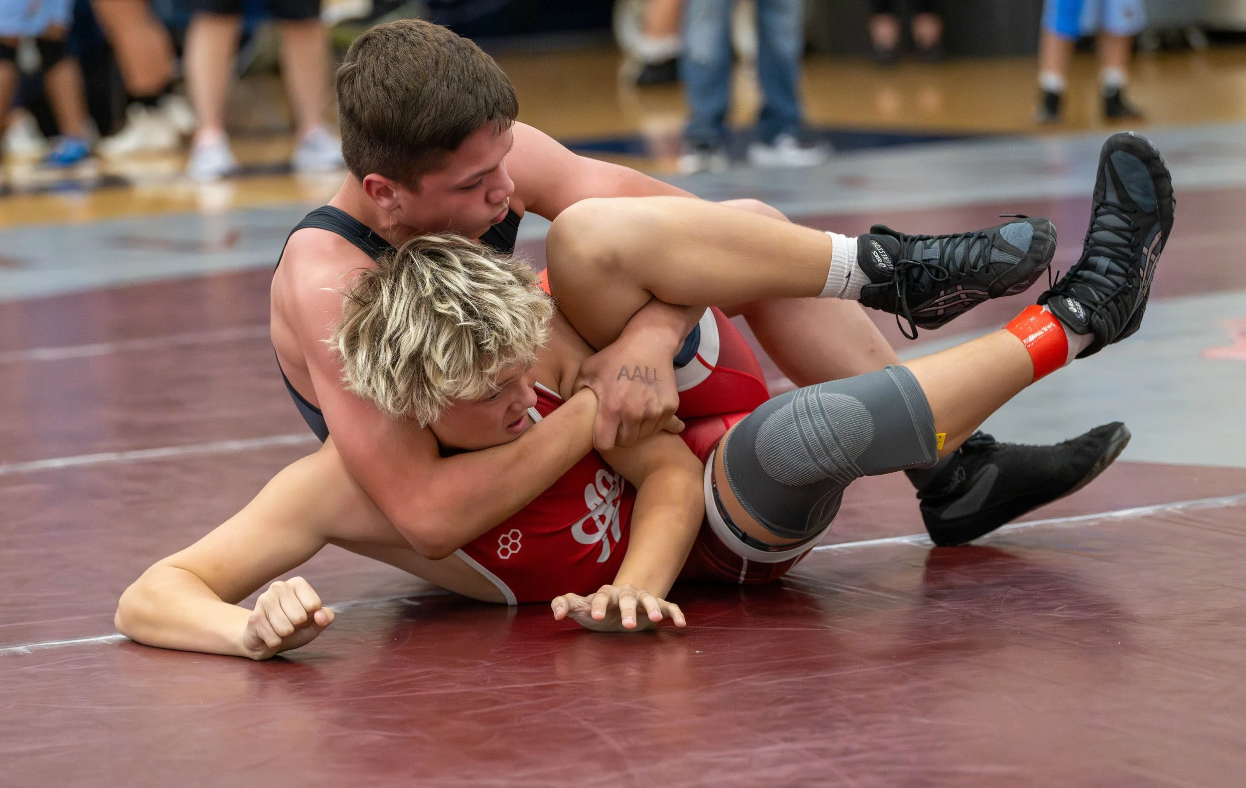 Two male wrestlers compete on a red wrestling mat, one in black gear and the other in red, inside a gymnasium with spectators in the background.