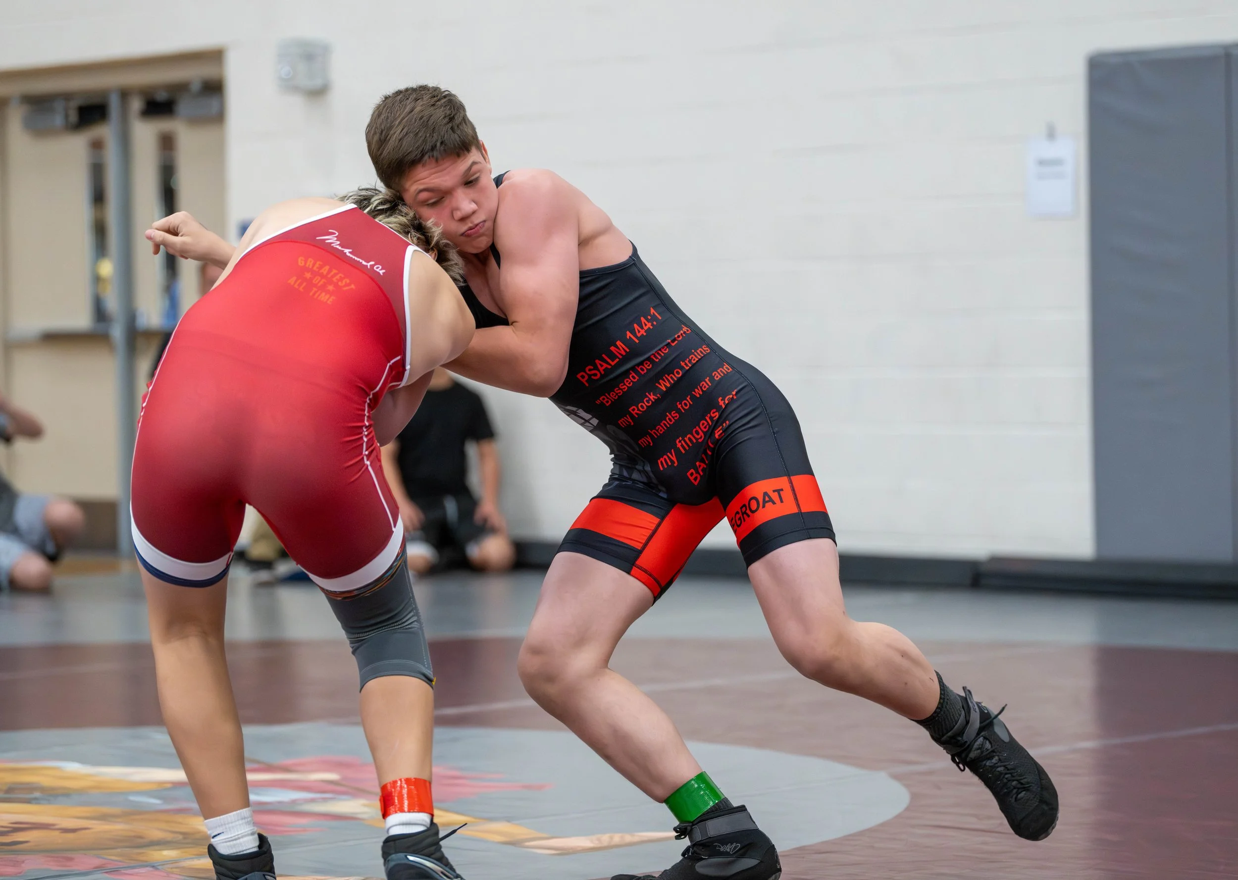 Two young male wrestlers wearing singlets engaged in a wrestling match on a mat indoors, with spectators sitting in the background.