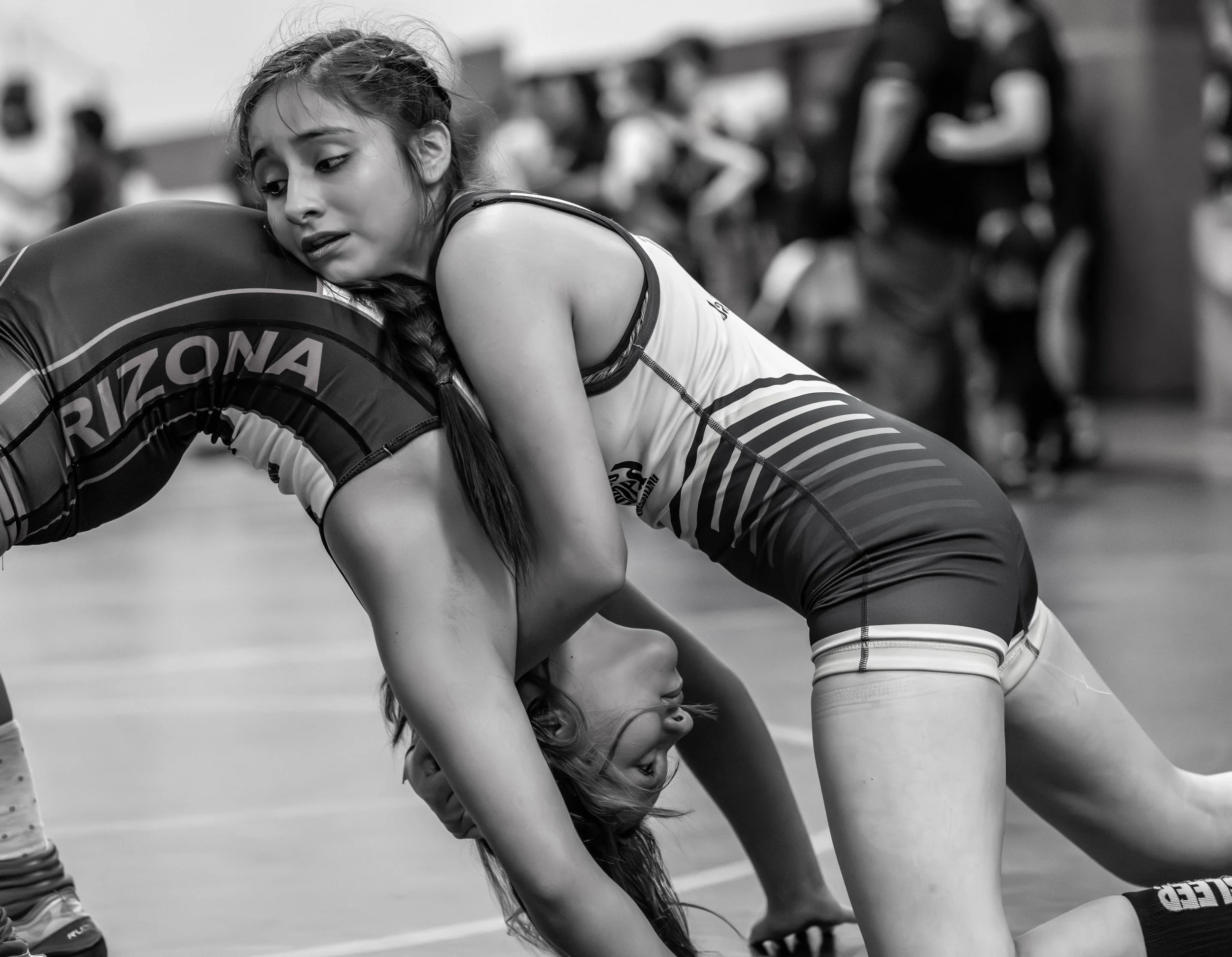 Two female wrestlers competing on the mat during a match, with one on top and the other on the bottom, in a gymnasium.