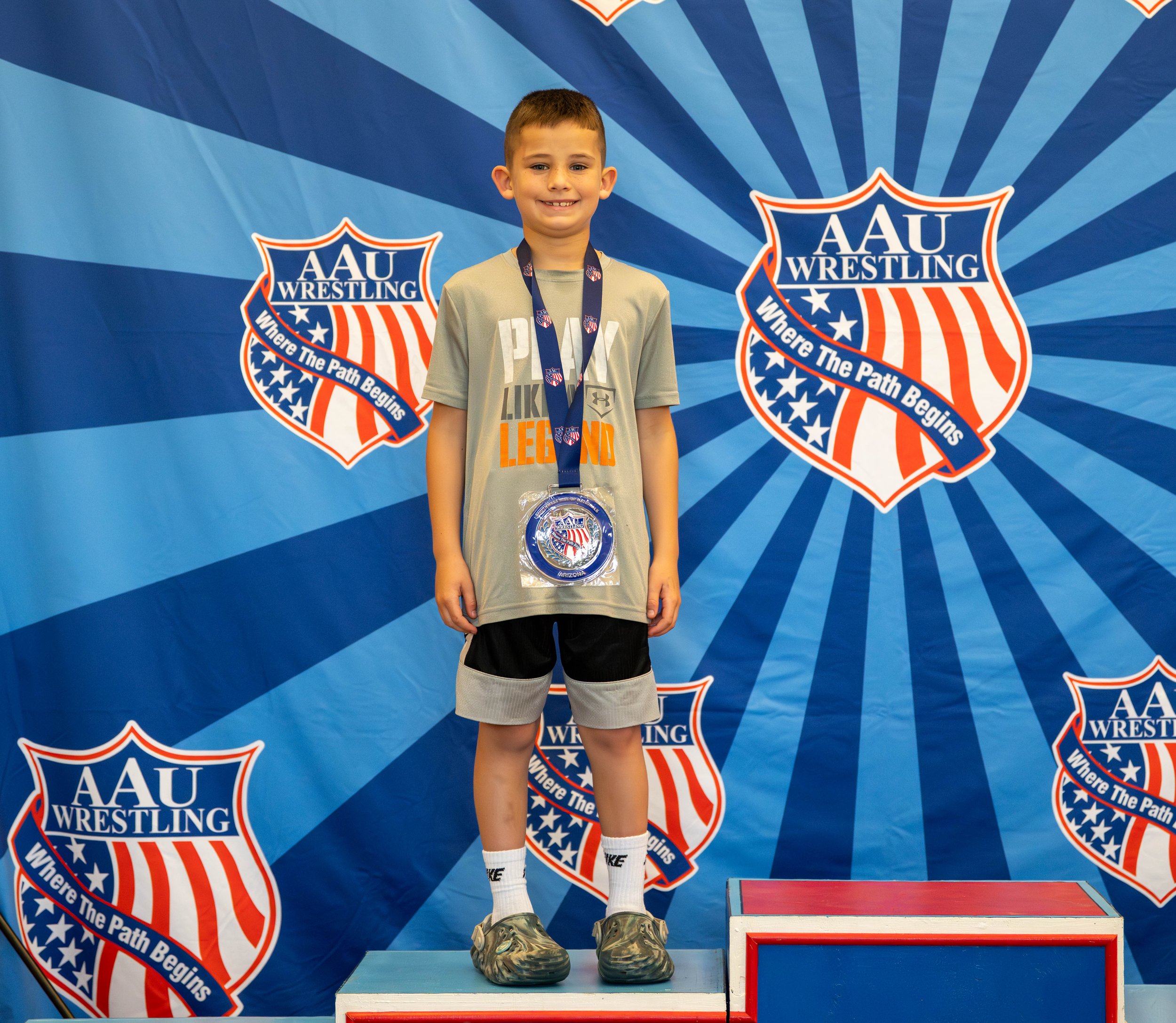 Young boy standing on a winners' podium with a medal around his neck, at an AAU Wrestling event. He is smiling, wearing a gray T-shirt, black shorts, and silver sneakers, with a background featuring the AAU Wrestling logo and the slogan "Where The Pa