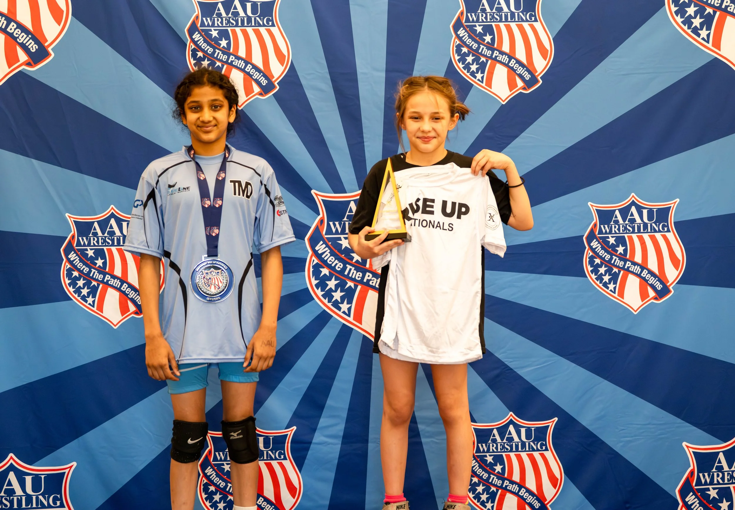 Two young female wrestlers stand on a blue victory podium in front of a backdrop with the AAU Wrestling logo. The girl on the left wears a light blue wrestling uniform with a medal around her neck, and the girl on the right holds a trophy and a white