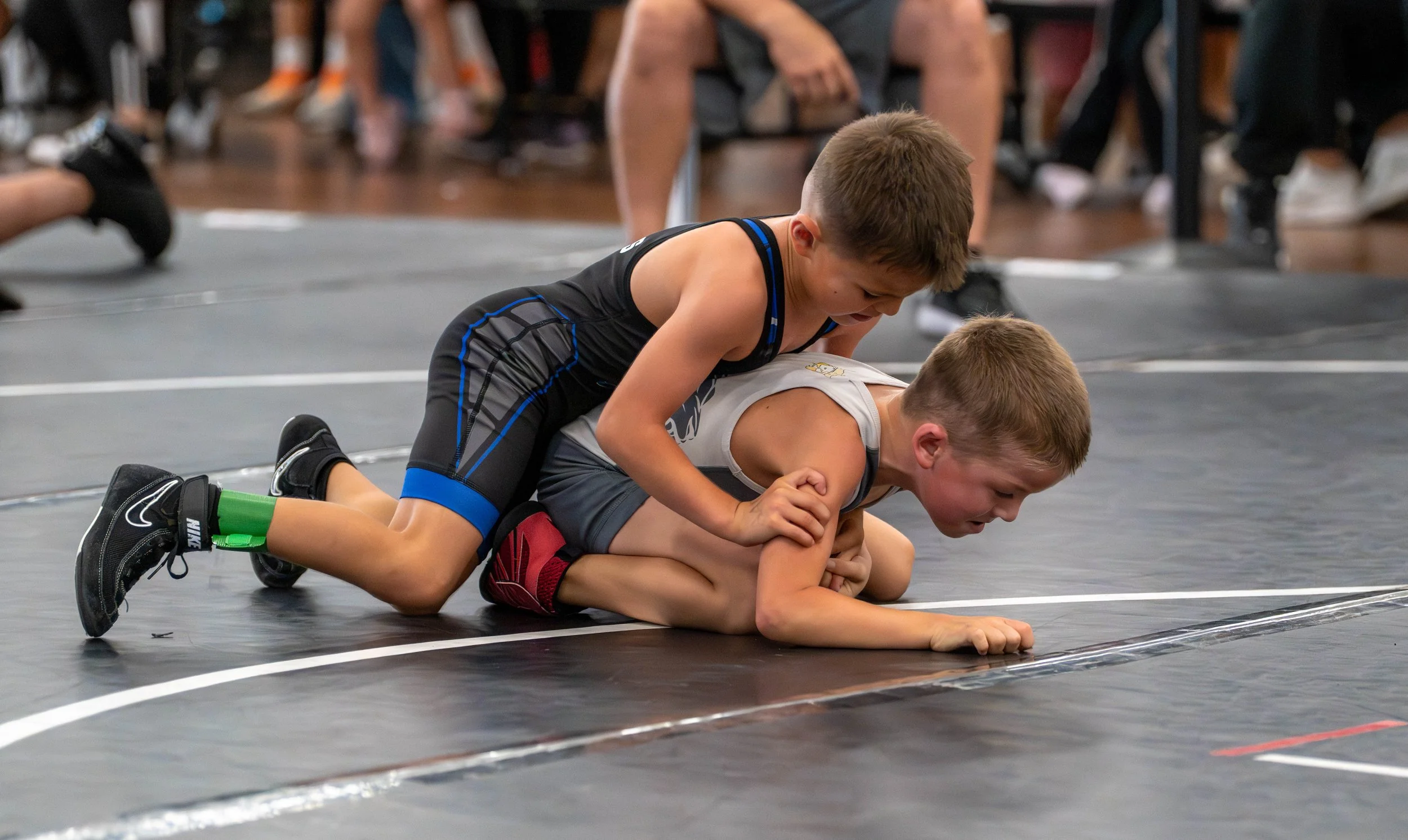 Two young boys wrestling on a black mat during a wrestling match, with the boy in black on top and the boy in gray on the bottom, onlookers in the background.