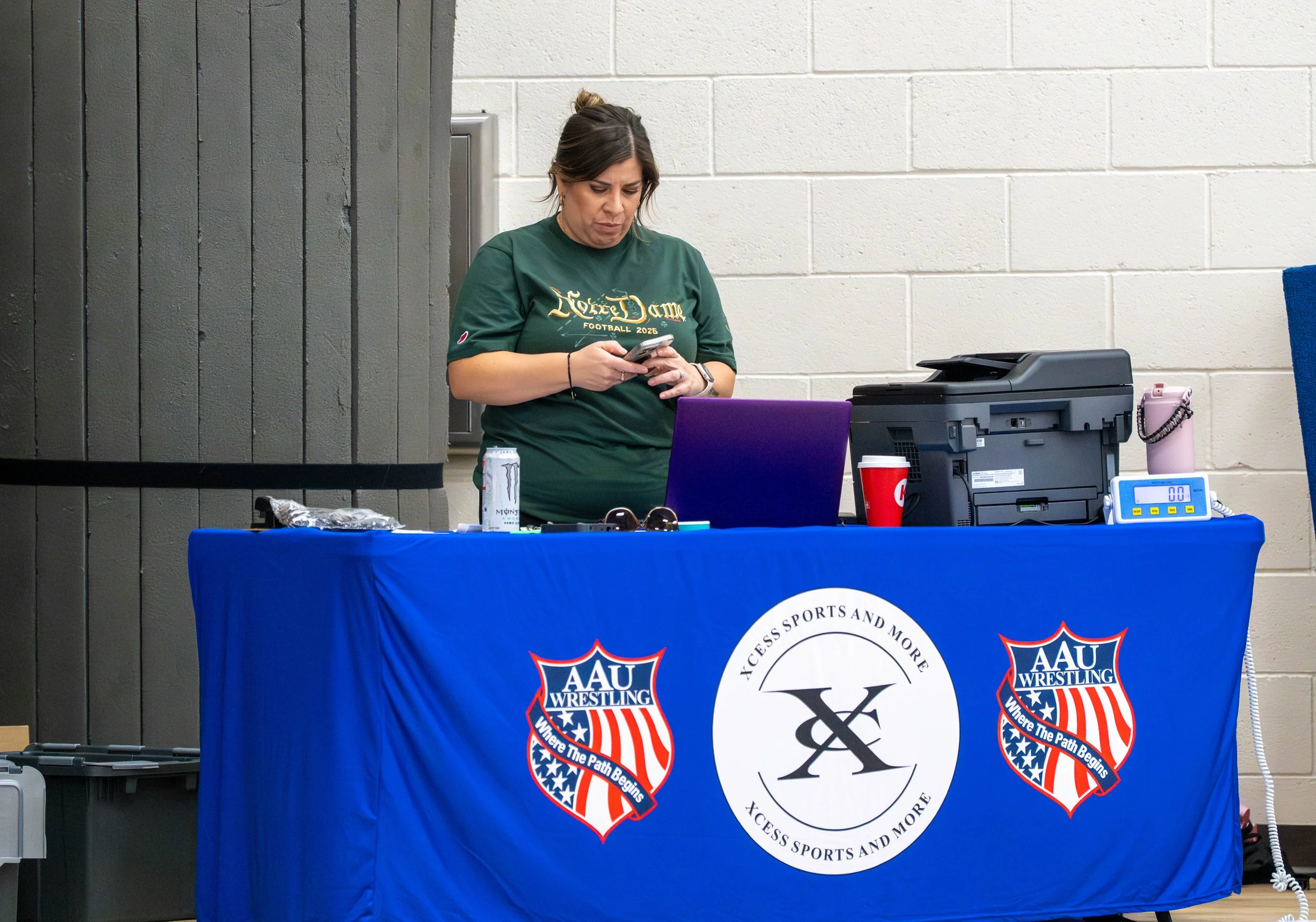 A woman in a green shirt standing behind a blue table with logos for AAU Wrestling and XCESS SPORTS AND MORE. She is looking at her phone. The table has a laptop, a printer, a red cup, and a pink water bottle.