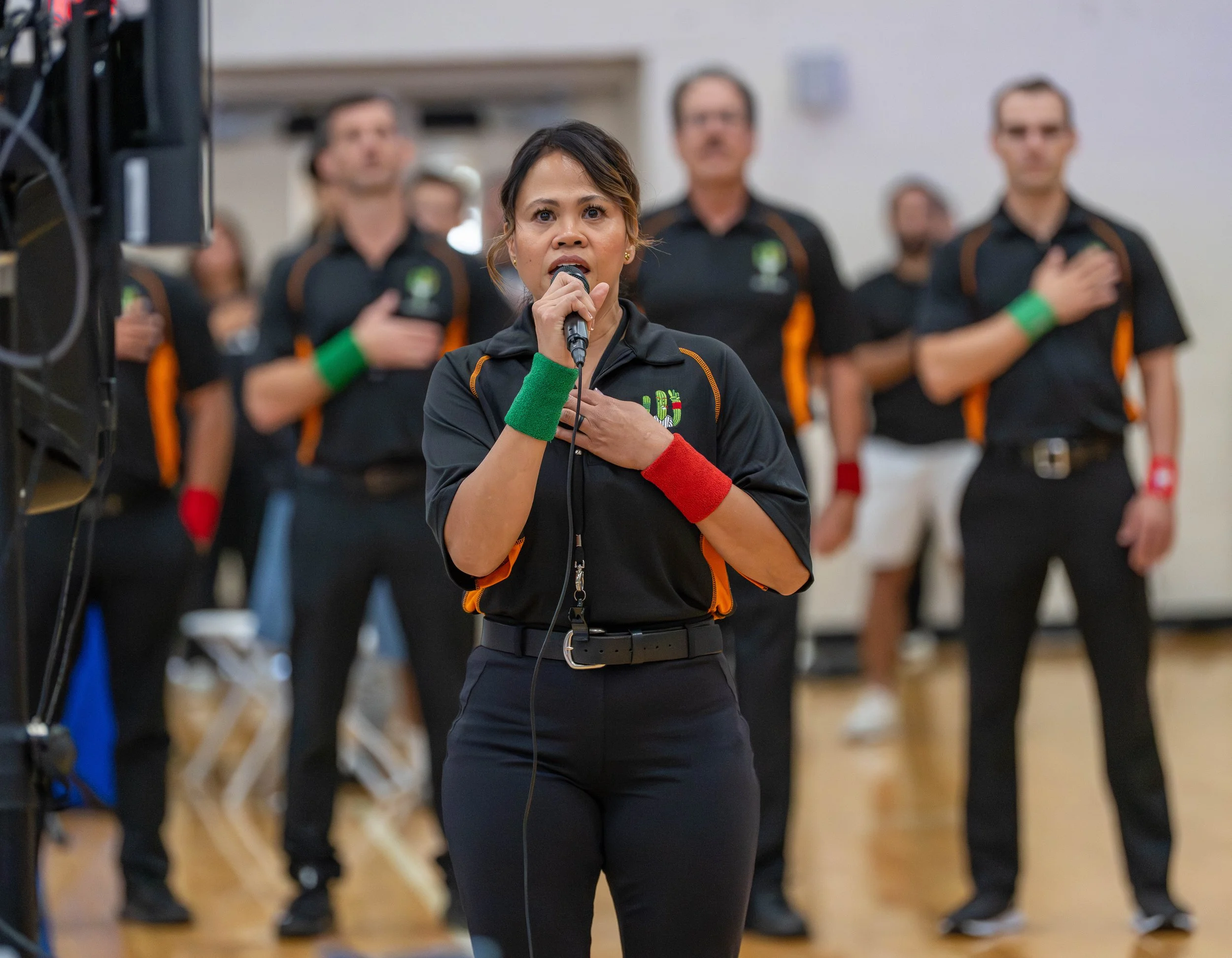 A woman in black sports attire with colorful wristbands, holding a microphone and placing her right hand on her chest while singing or speaking. Behind her are several men also in sports attire, with their right hands on their chests, standing in a g