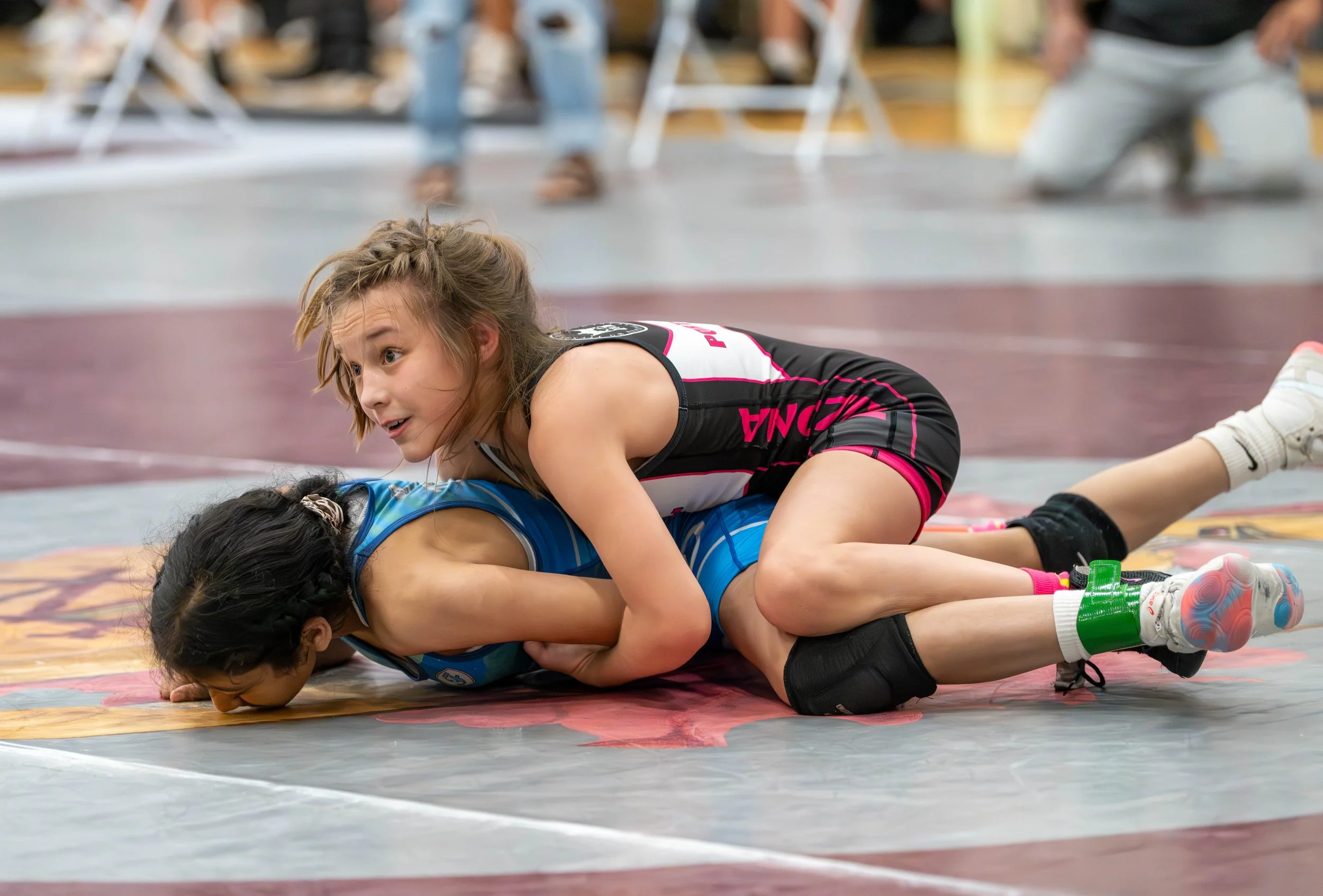Two young female wrestlers in action on a wrestling mat during a match, with one girl in a blue uniform on the ground and the other girl in a black and pink uniform on top, attempting a move.