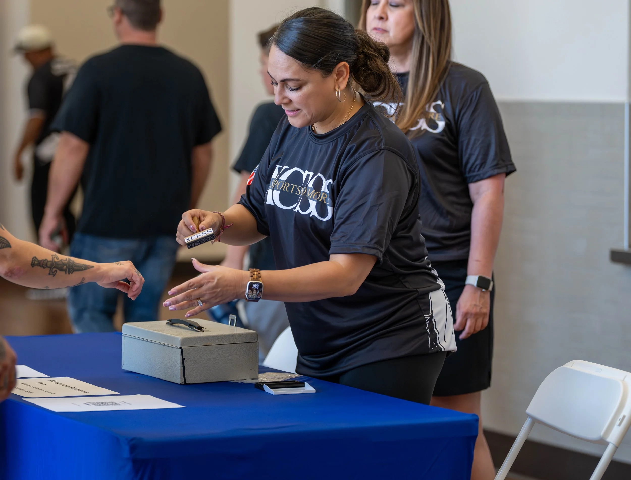 A woman wearing a black sports jersey places a bracelet on another person's wrist at a registration table during an event. There are other people in the background.