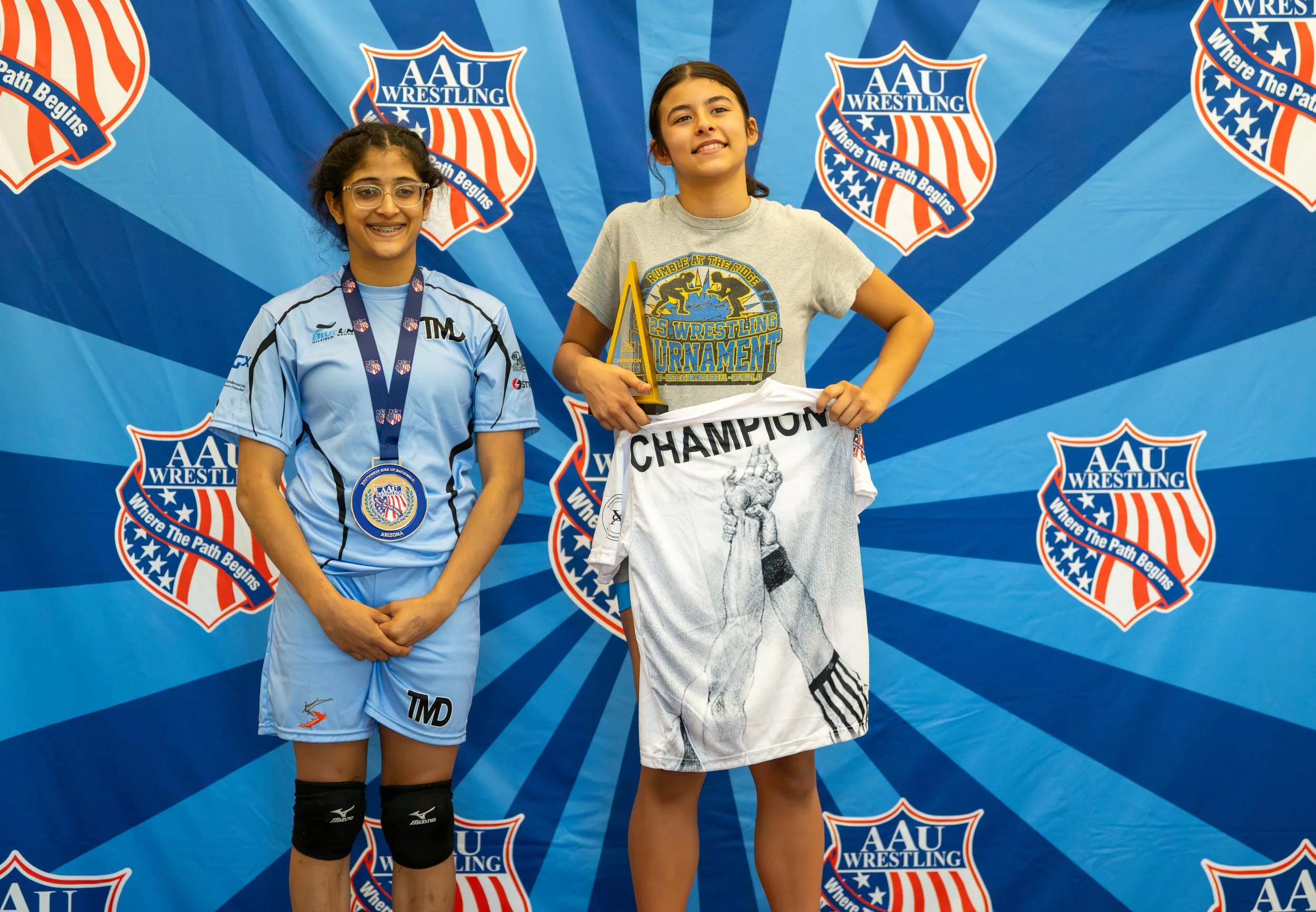 Two young girls standing on a victory podium at an AAU Wrestling event. The girl on the left wears a blue wrestling uniform with a medal around her neck and black knee pads. The girl on the right holds a champion towel and a trophy, wearing a gray t-