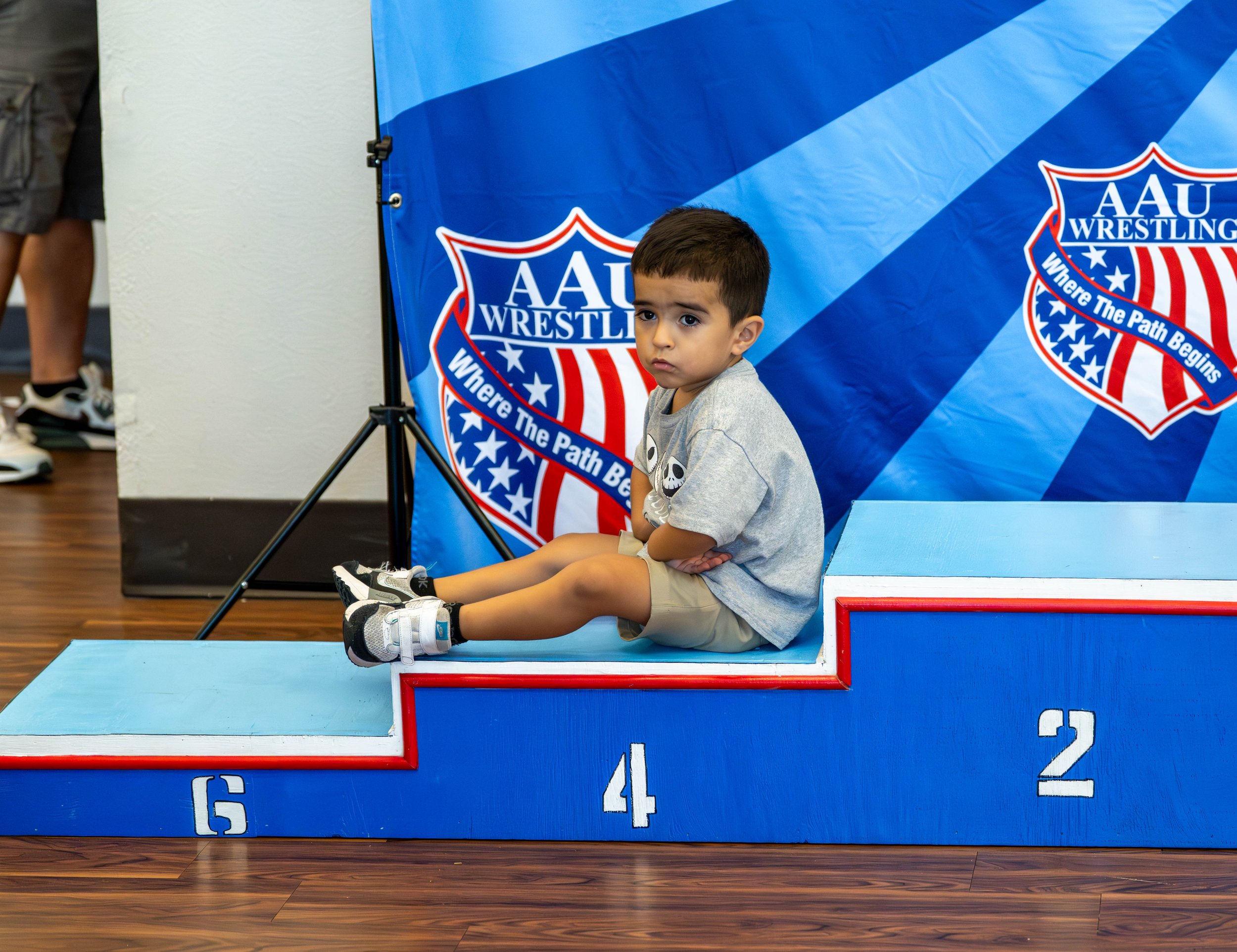 A young boy sitting on a 4th place winner's podium at an AAU wrestling event, with a large blue AAU wrestling banner in the background.