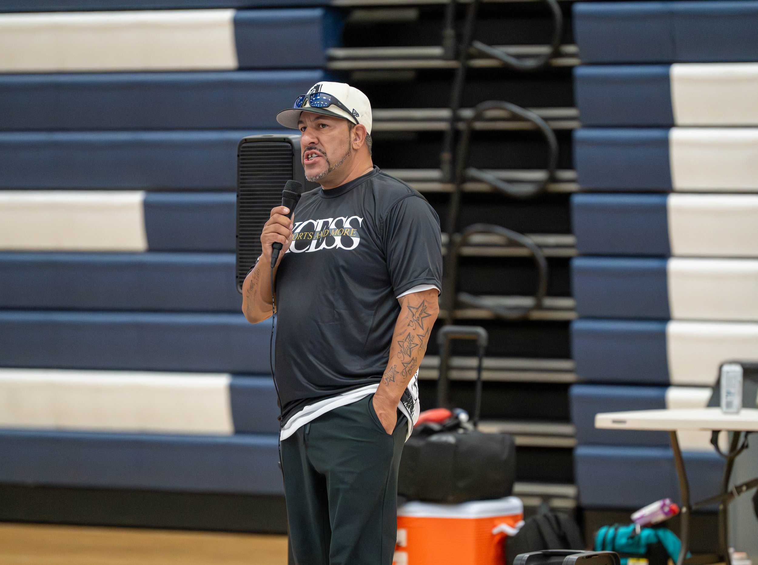 A man wearing a blackSport shirt, sunglasses, and a white cap is speaking into a microphone in a gymnasium with blue and white bleachers in the background. There is a table with a cooler and other equipment beside him.