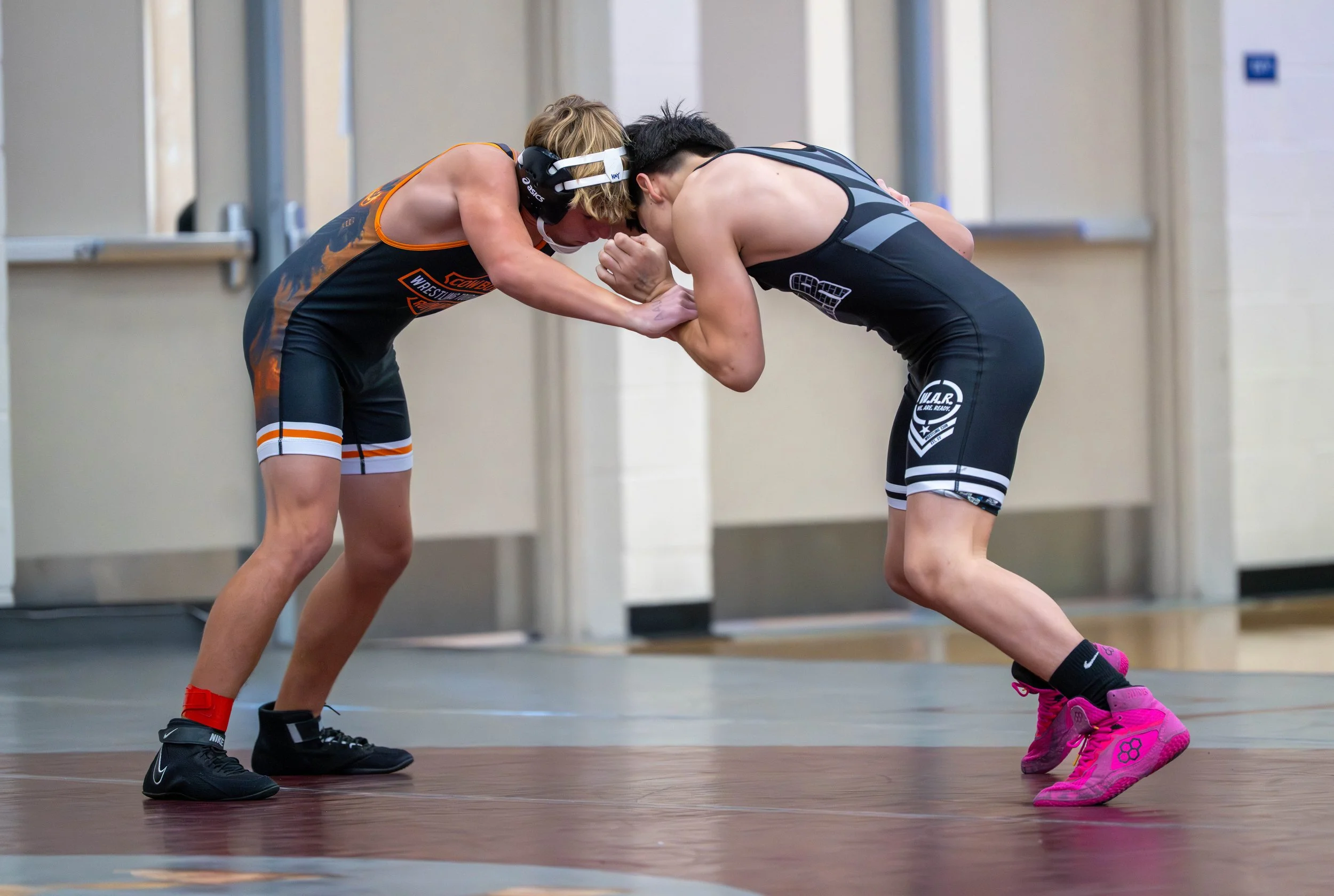 Two young wrestlers grappling on a mat during a match, one in black and pink shoes with pink shoes, the other in black shoes with white stripes, both dressed in athletic singlets.
