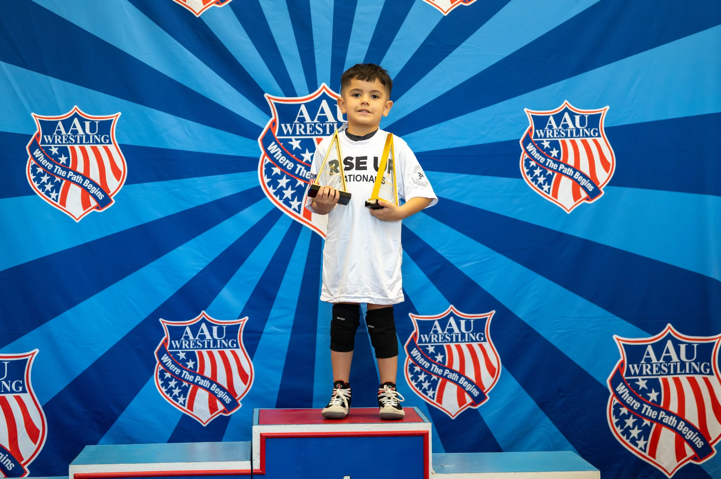 Young boy standing on a winners' podium holding two trophies at an AAU wrestling event, with multiple AAU Wrestling banners in the background.