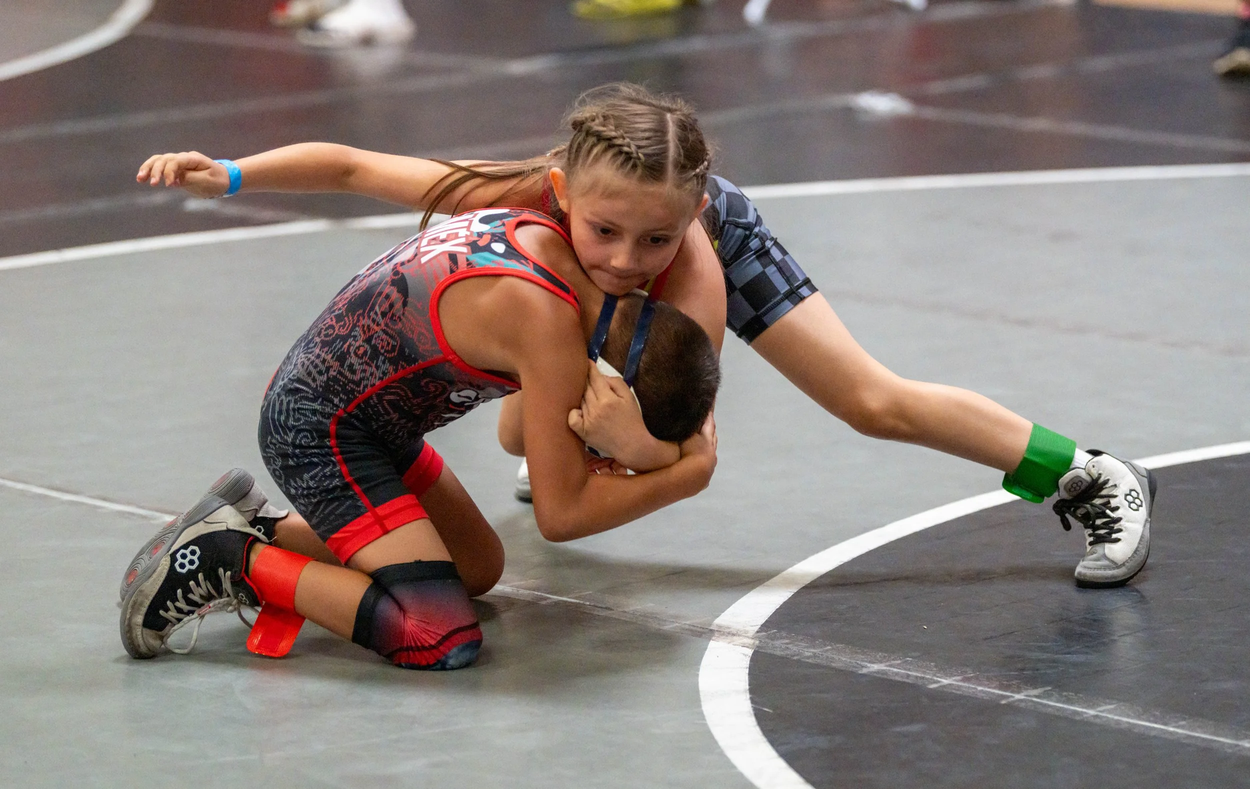 Two young boys wrestling on a gymnasium mat, one in red and the other in blue, with the boy in red on his knees and the boy in blue grabbing his head.