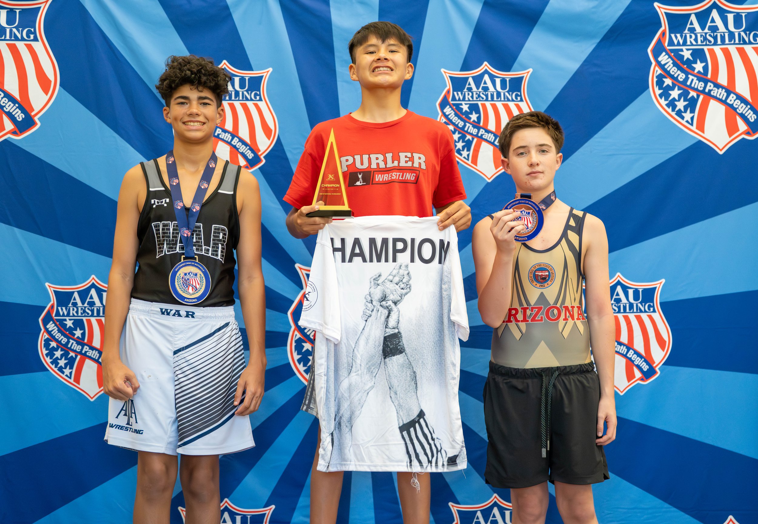 Three young boys on a winners' podium at a wrestling event, holding medals and a trophy, with a blue background displaying the AAU Wrestling logo.