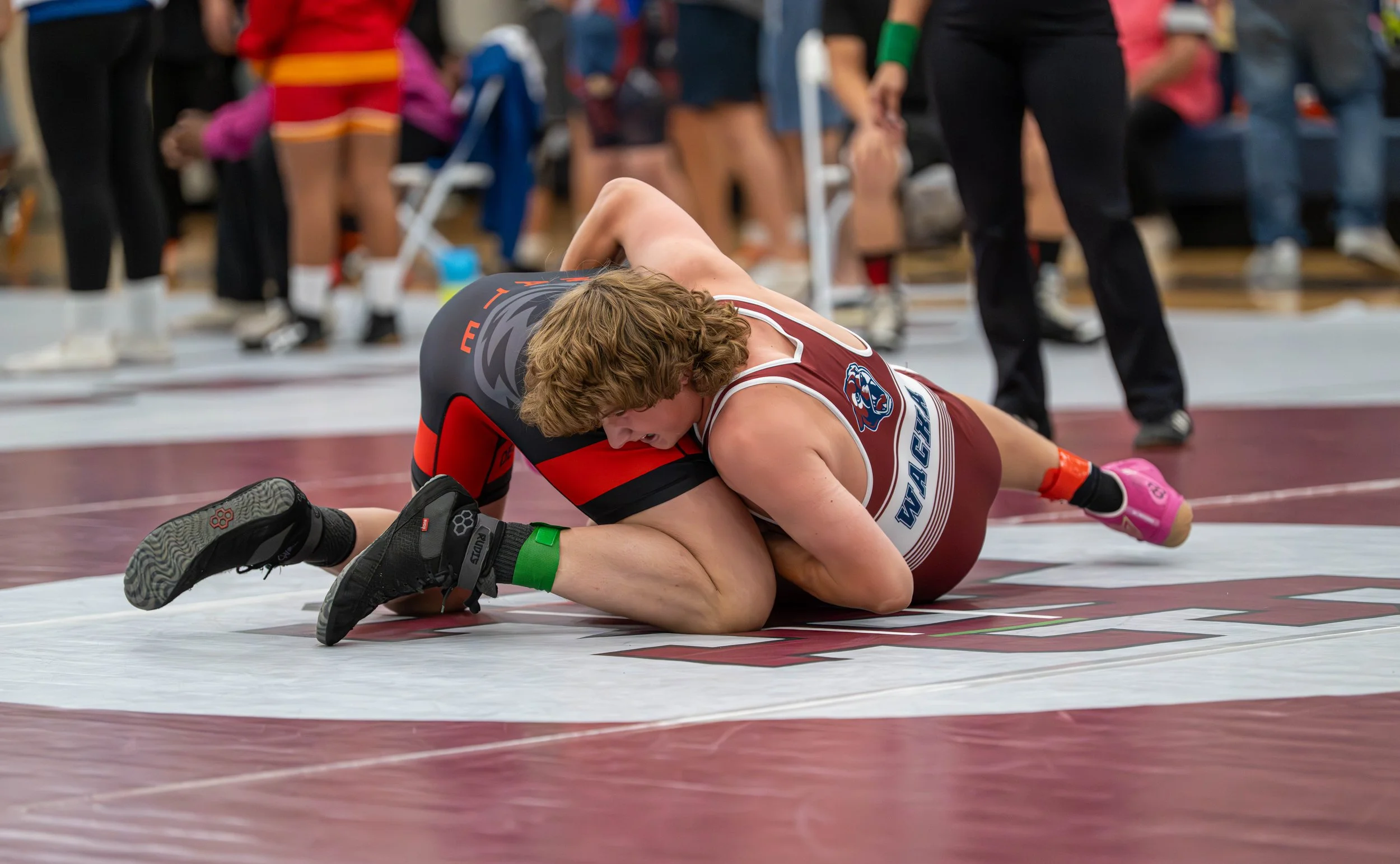 Two wrestlers competing on a wrestling mat, with one in a maroon singlet and the other in a black and red singlet, engaged in a match during a sporting event.