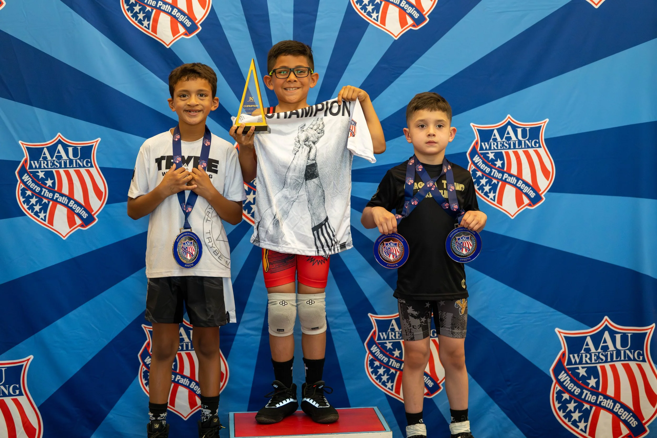 Three young boys standing on a winners' podium at an AAU Wrestling event with a blue backdrop displaying the AAU Wrestling logo. The boy in the center, holding a trophy and wearing a white shirt with the word "Champion," is on the highest podium step