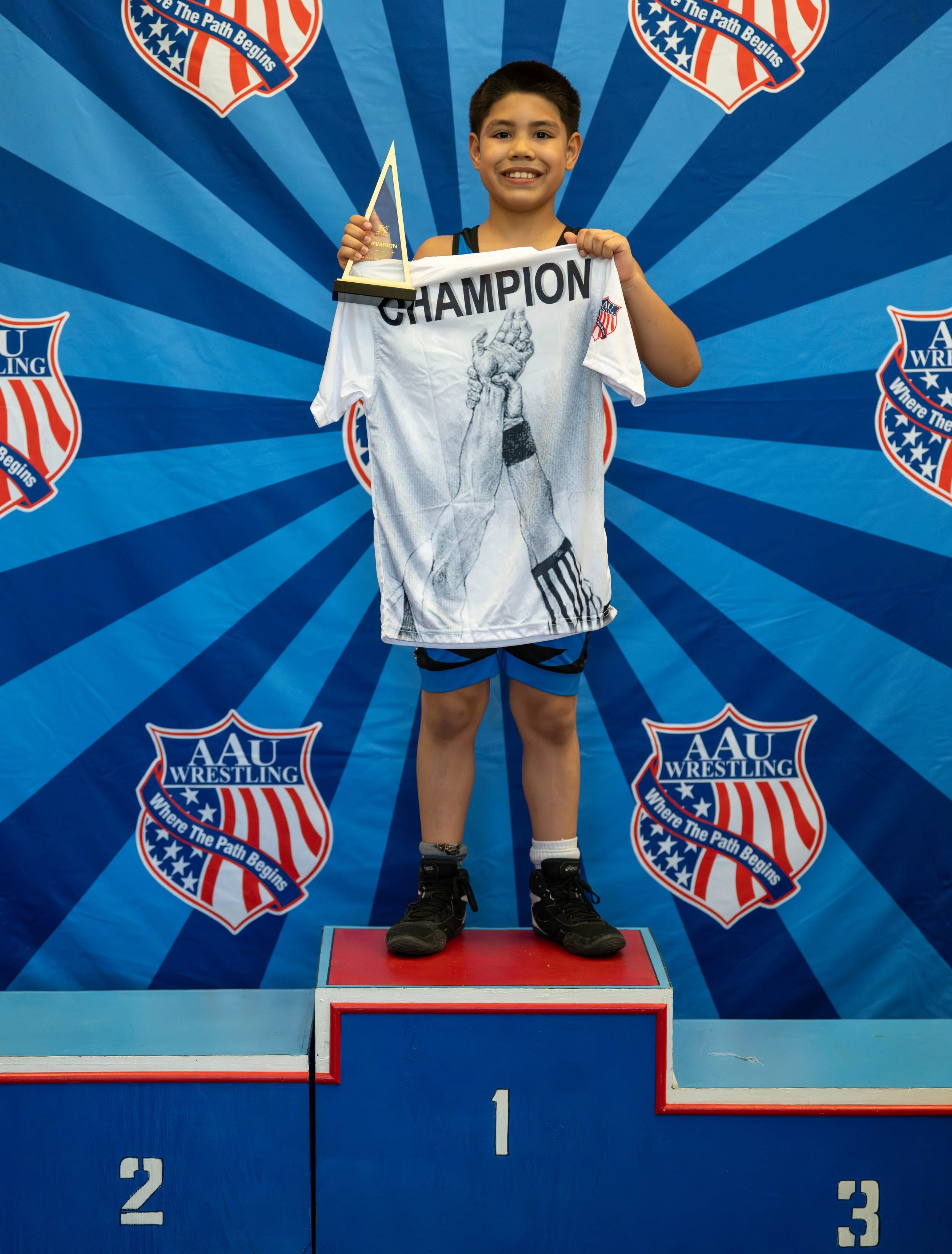 A young boy standing on the first place podium holding a trophy and a shirt that says "CHAMPION." He is smiling and wearing wrestling gear, with a banner of AAU Wrestling in the background.