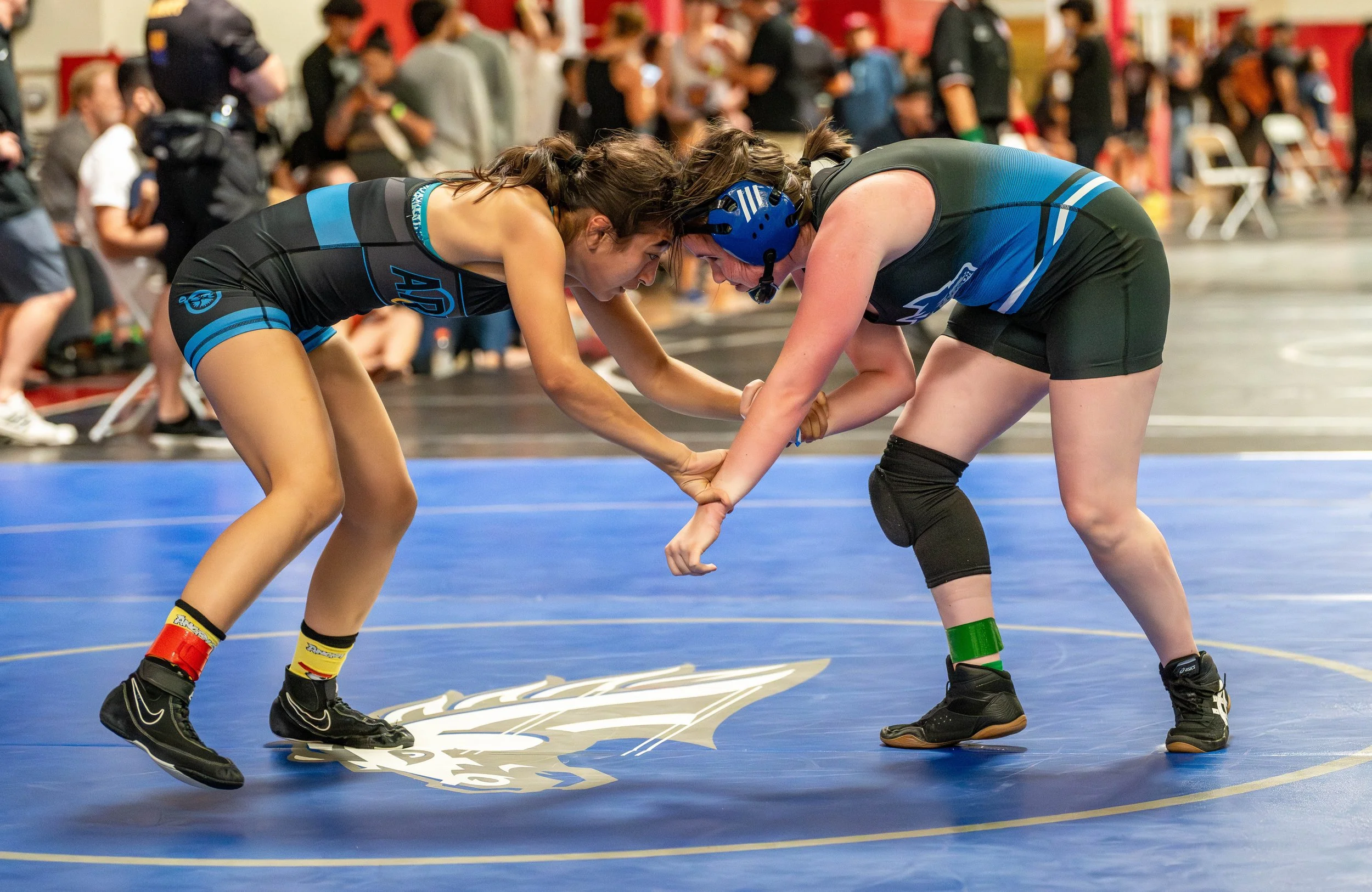 Two female wrestlers in black and blue singlets engaged in a wrestling match on a blue mat, with a crowd and officials in the background.