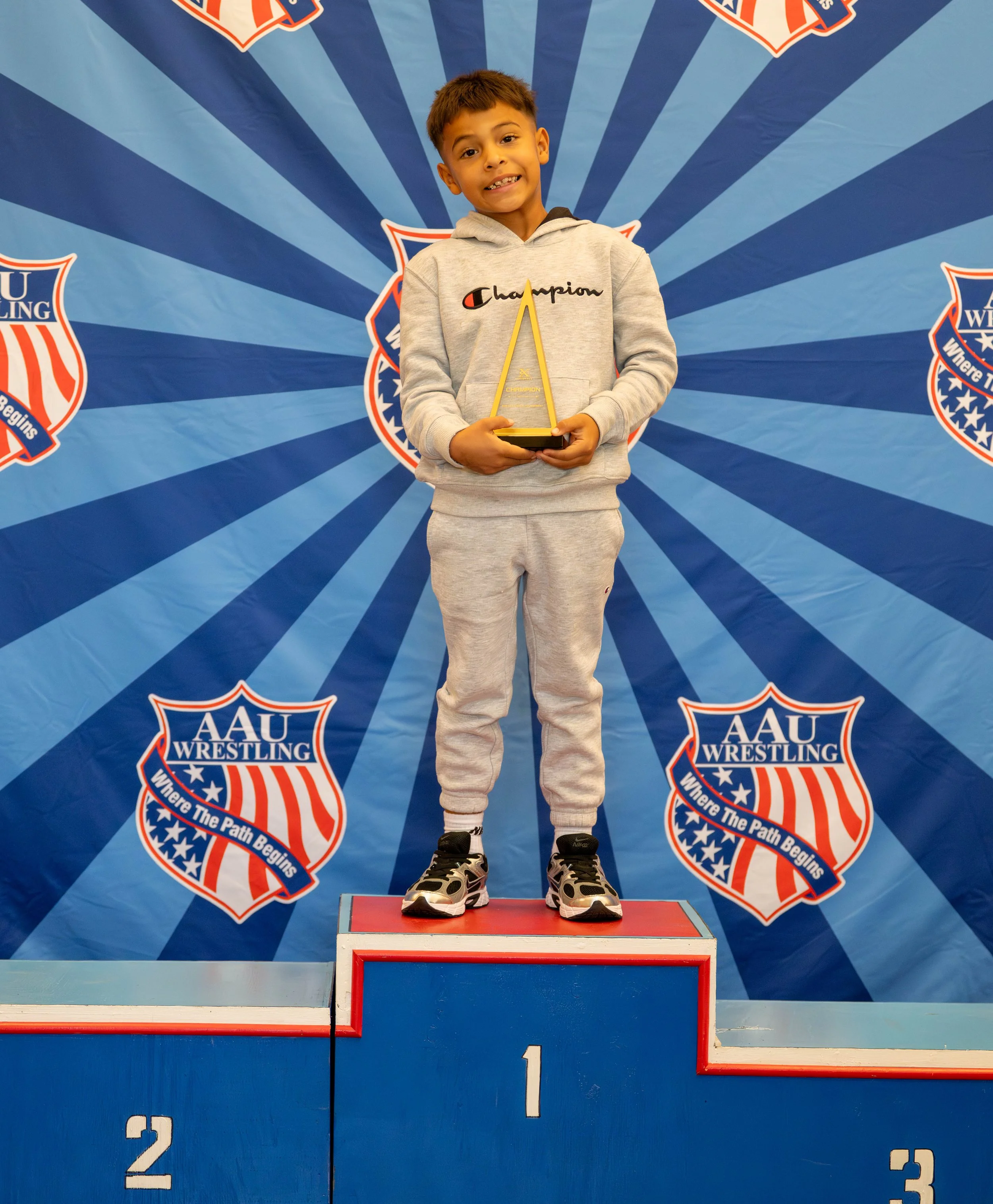 Young boy standing on first place podium holding a trophy. Background features AAU Wrestling banners with the slogan 'Where The Path Begins'.