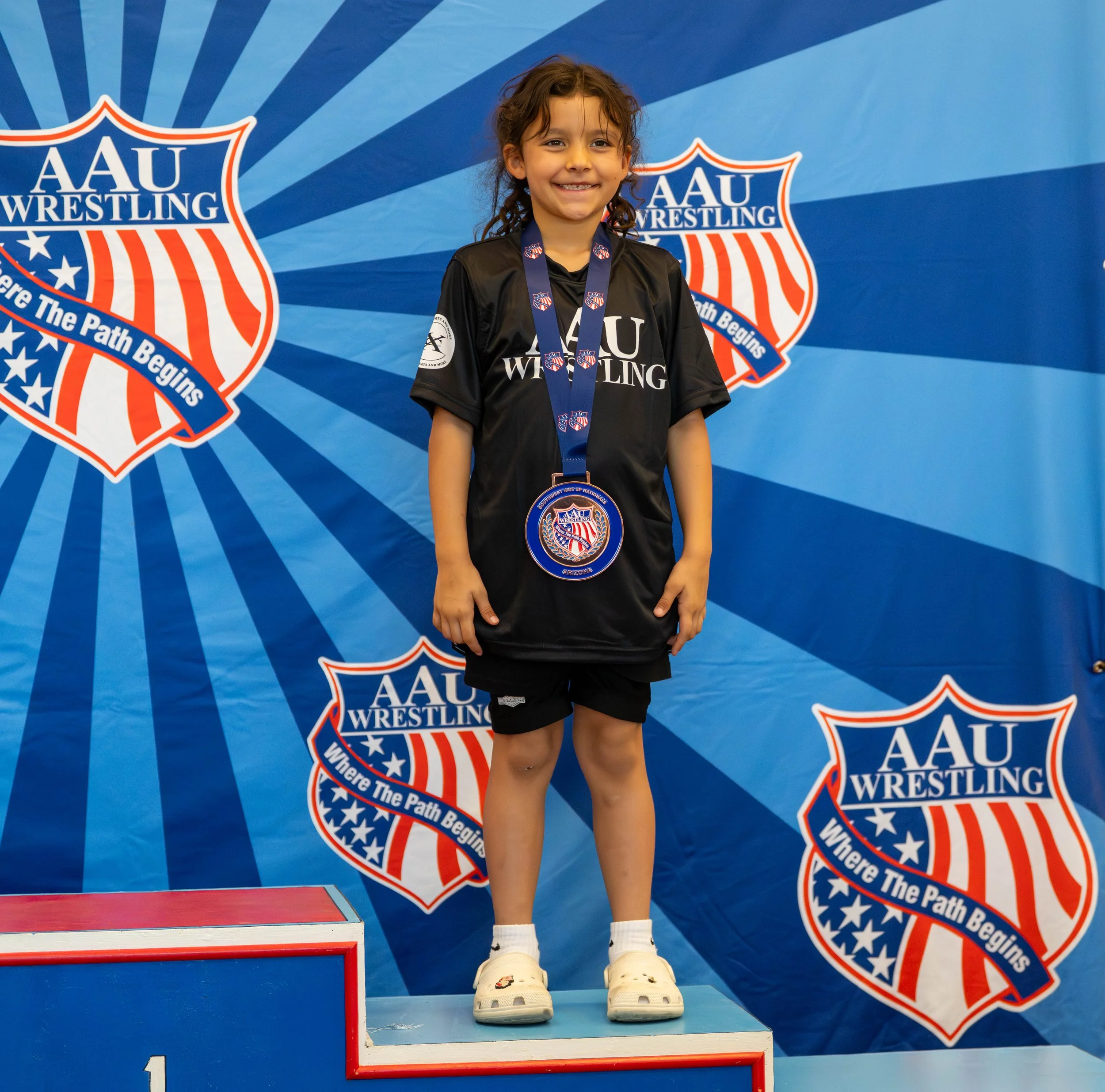 Young girl standing on a winners' podium at an AAU wrestling event, wearing a medal and a black AAU wrestling shirt, in front of a blue background with AAU wrestling logos.