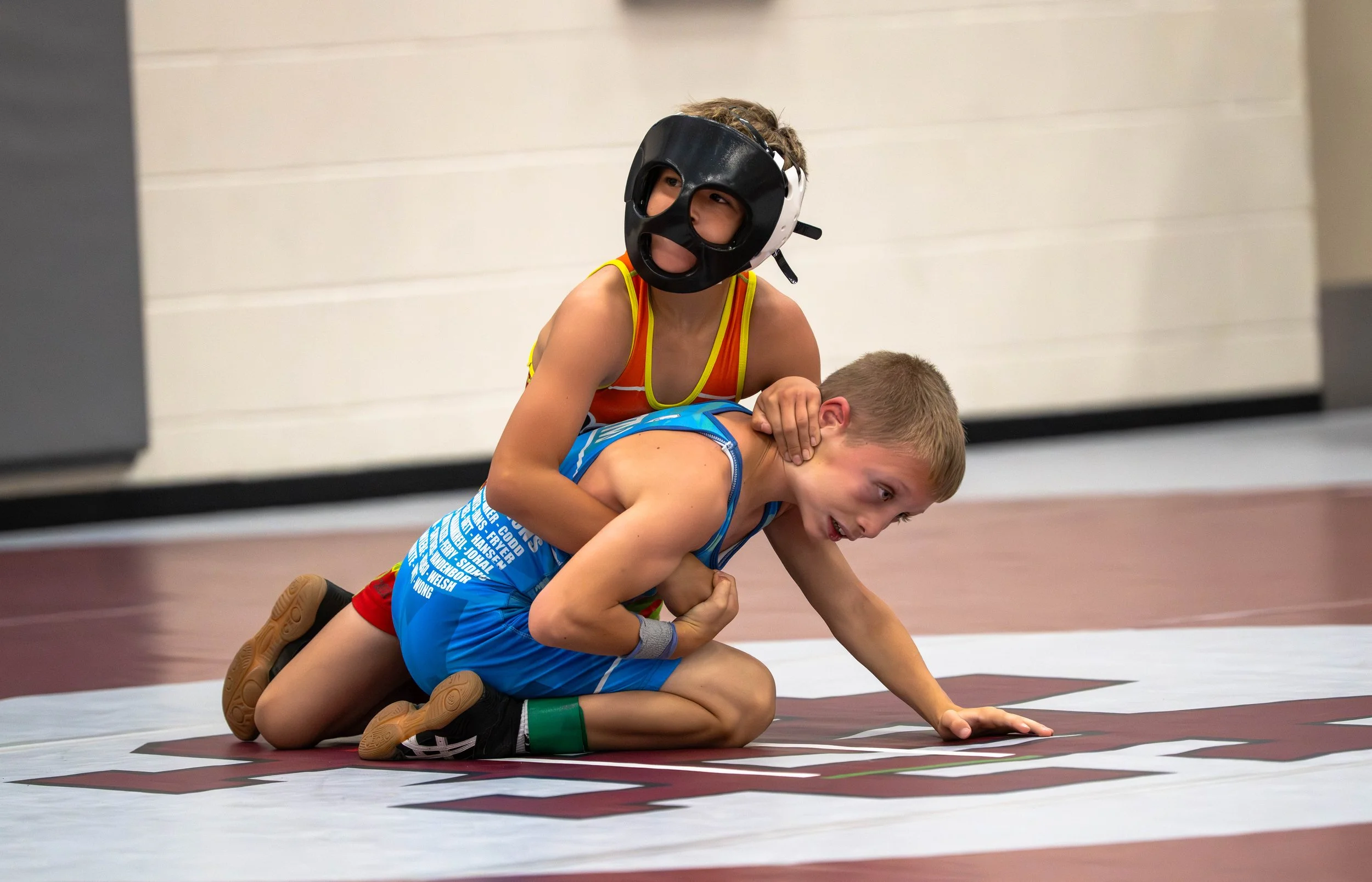 Young boy in a blue wrestling singlet being pinned by an opponent wearing a protective headgear in a wrestling match.