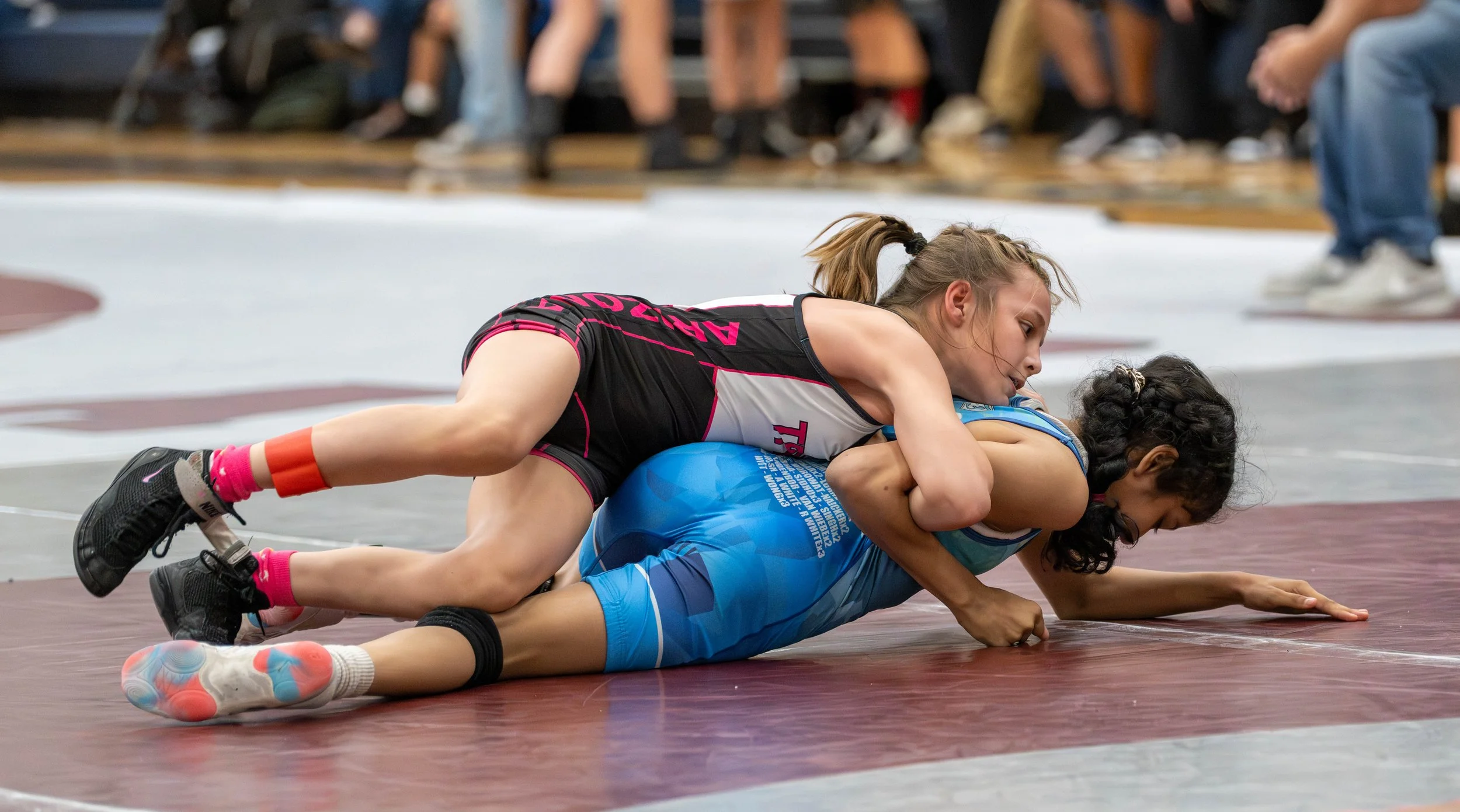 Two female wrestlers competing on a wrestling mat, one in a black and pink singlet and the other in a blue singlet, during a match.