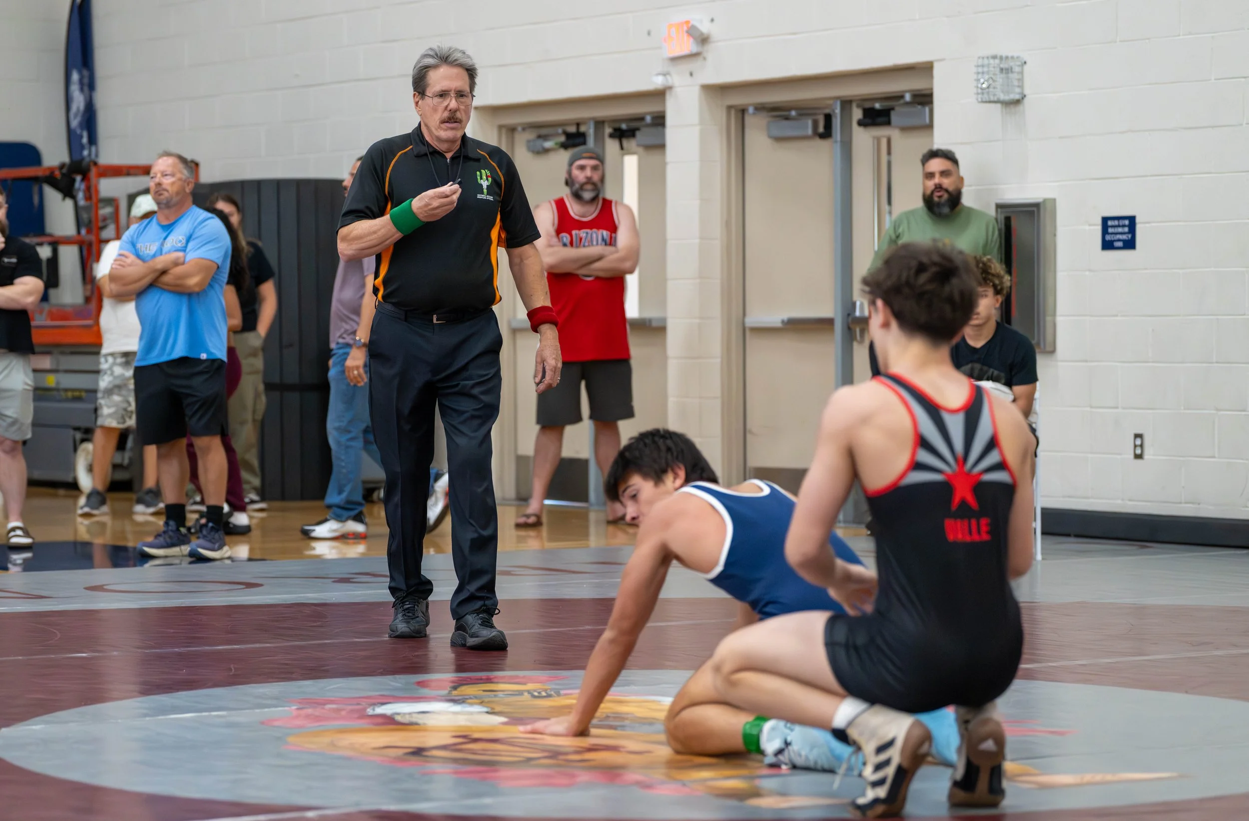 A wrestler in a blue singlet is down on the mat, appearing to be injured, while another wrestler in a black singlet with red accents kneels beside him. An official stands in front of them, holding a whistle, during a wrestling match in a gymnasium wi