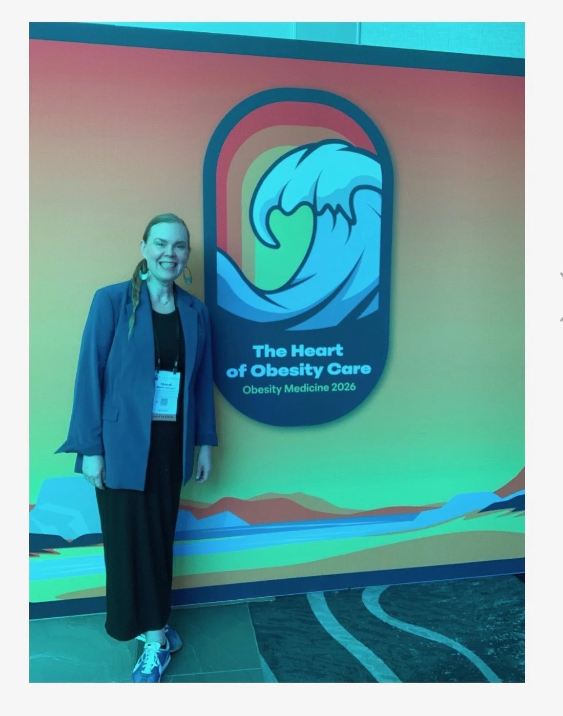 A woman standing next to a sign that reads 'The Heart of Obesity Care, Obesity Medicine 2026,' featuring a large stylized wave and rainbow design.